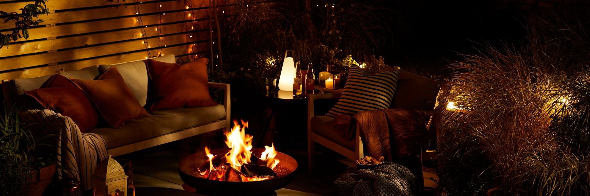 Night time scene in garden with cushioned chairs, candles and a lit fire pit