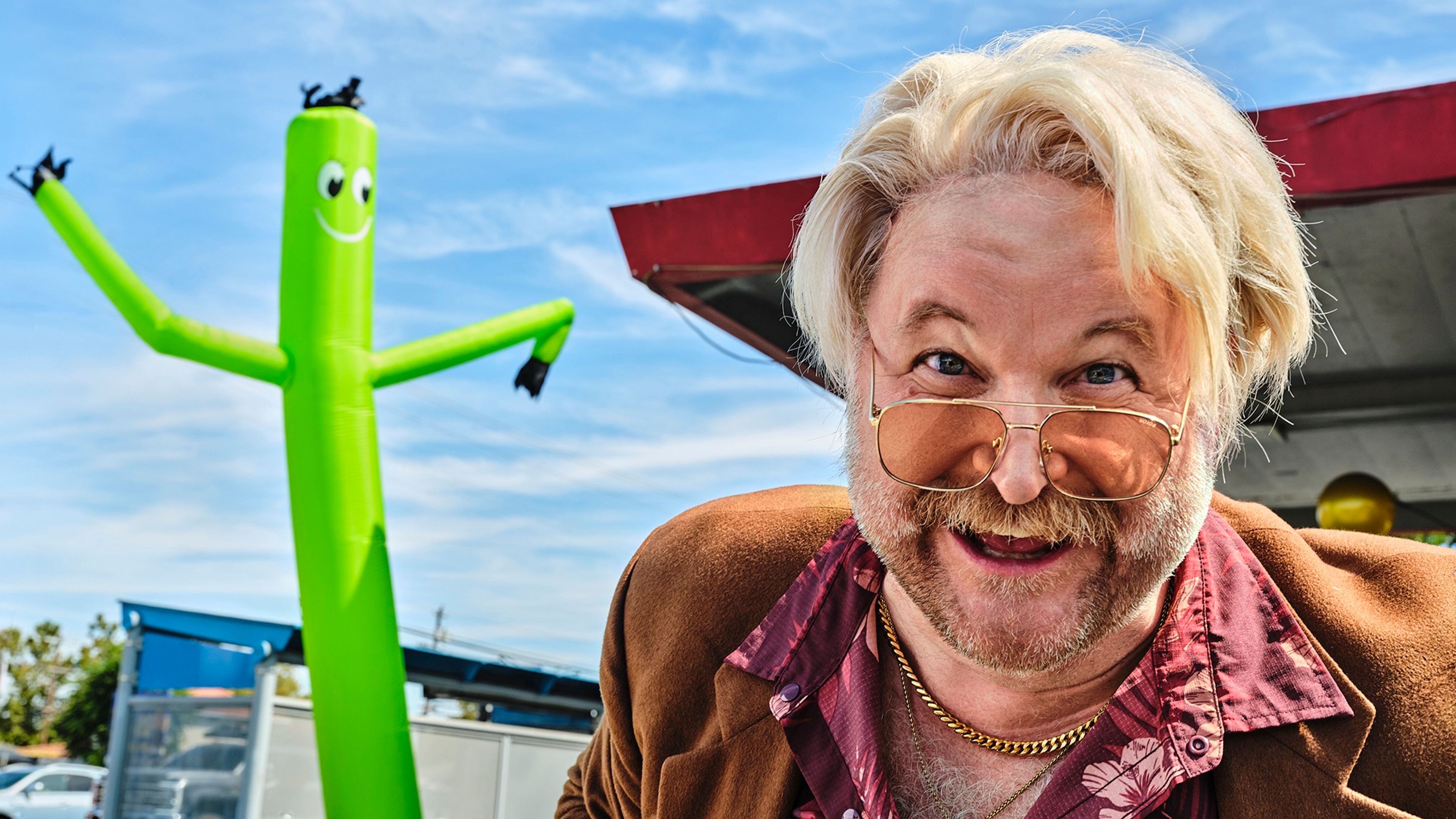 A man with blond hair and tinted glasses grins beside a green inflatable tube figure.