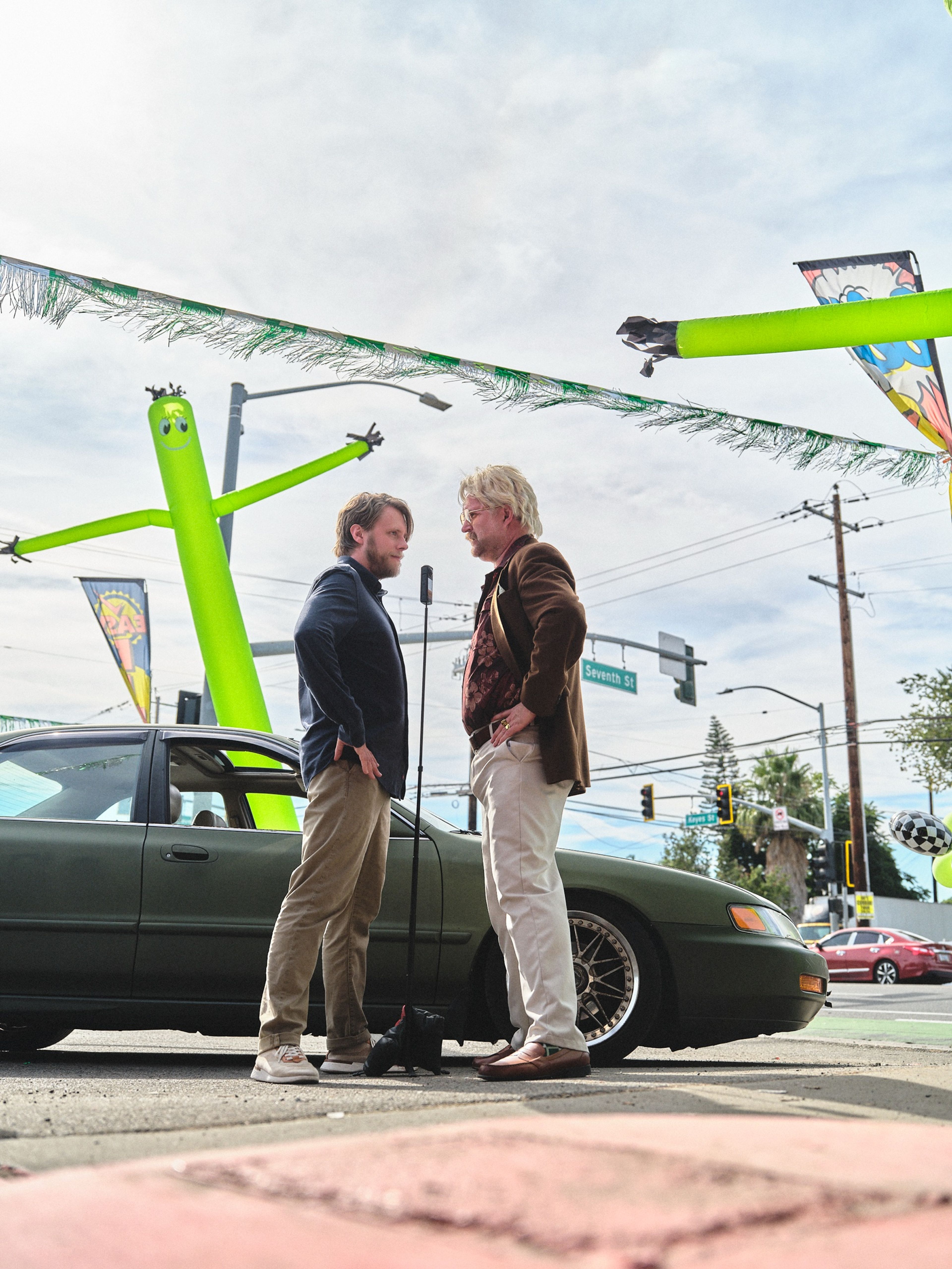 Low-angle shot of two actors facing off beside an olive-green sedan, with green inflatable tube figures and streamers against the sky.