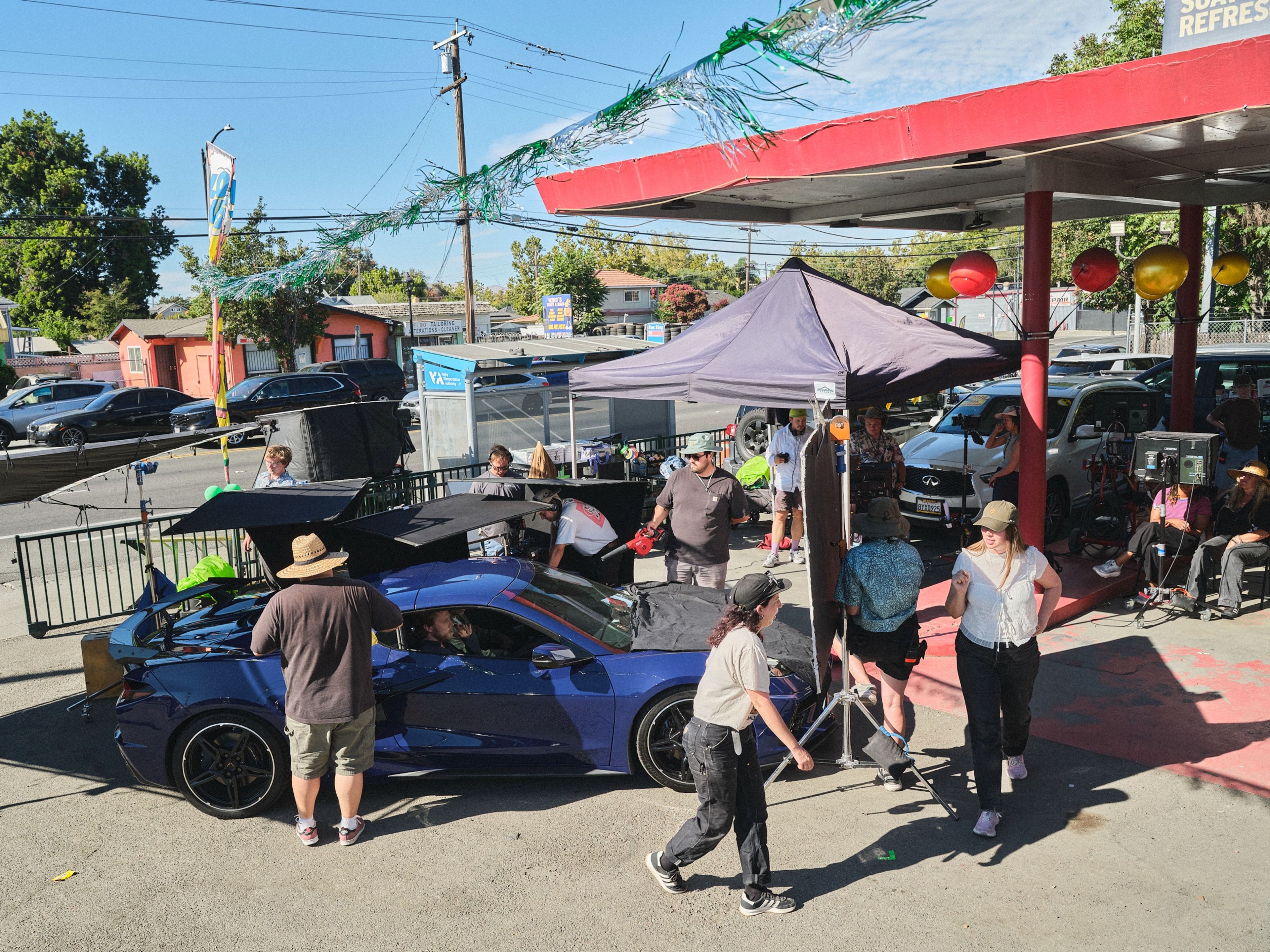 From above, a film crew works around a blue sports car under a red gas-station canopy, with a pop-up tent, lights and a row of crew in chairs at the monitors.