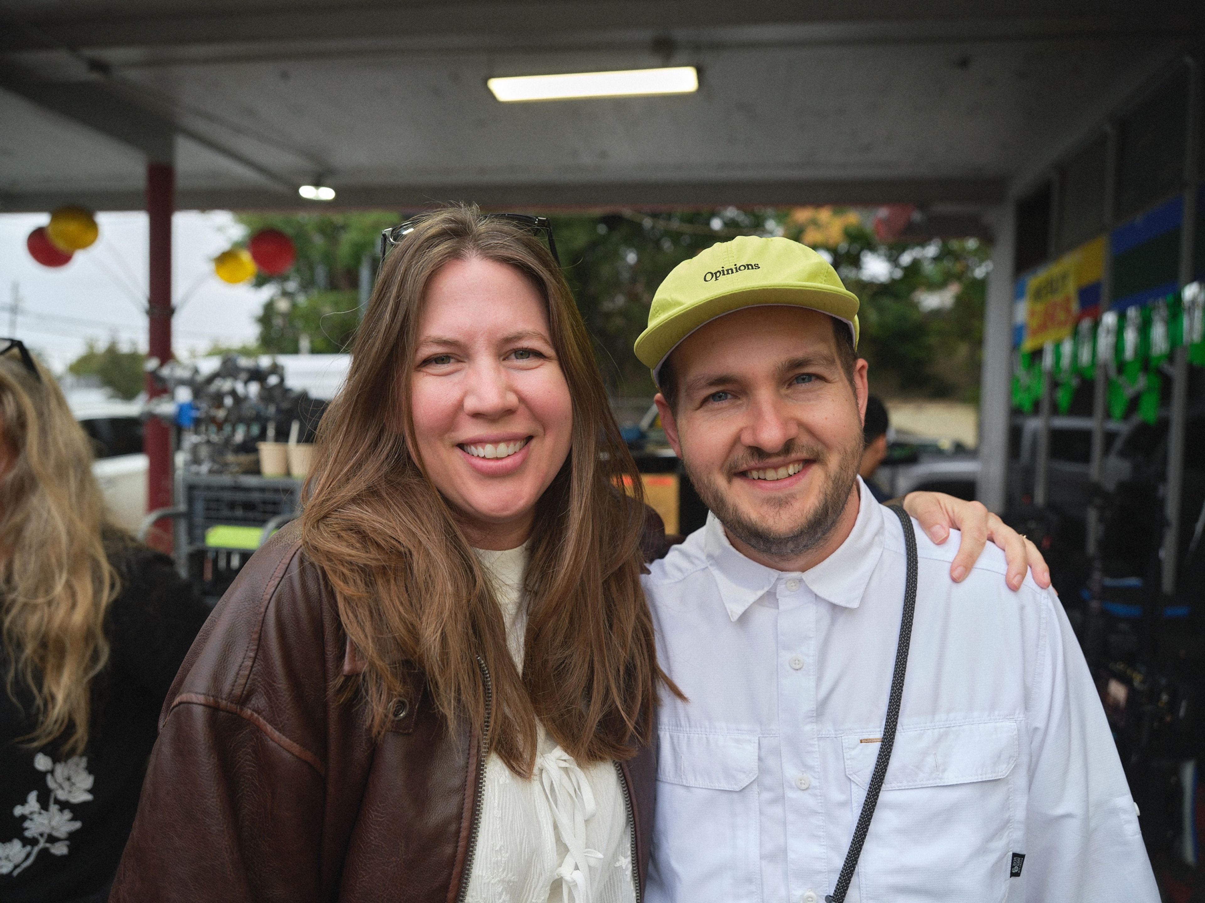 A woman in a leather jacket and a man in a yellow cap smile for the camera under the car-lot canopy.