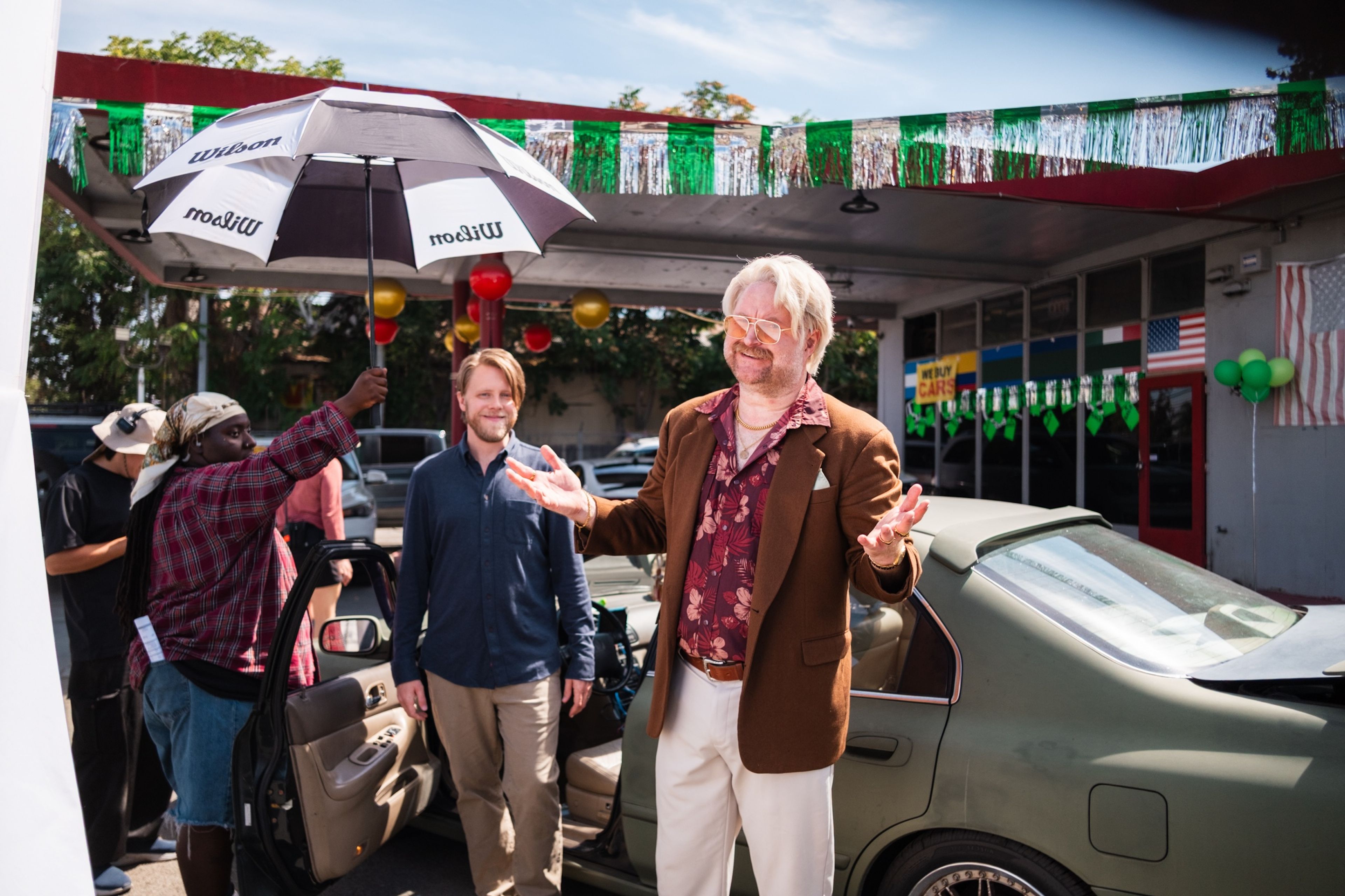 On set at a car lot hung with green and silver streamers, a man in a brown blazer and tropical-print shirt gestures with open arms while a crew member holds an umbrella over the scene.