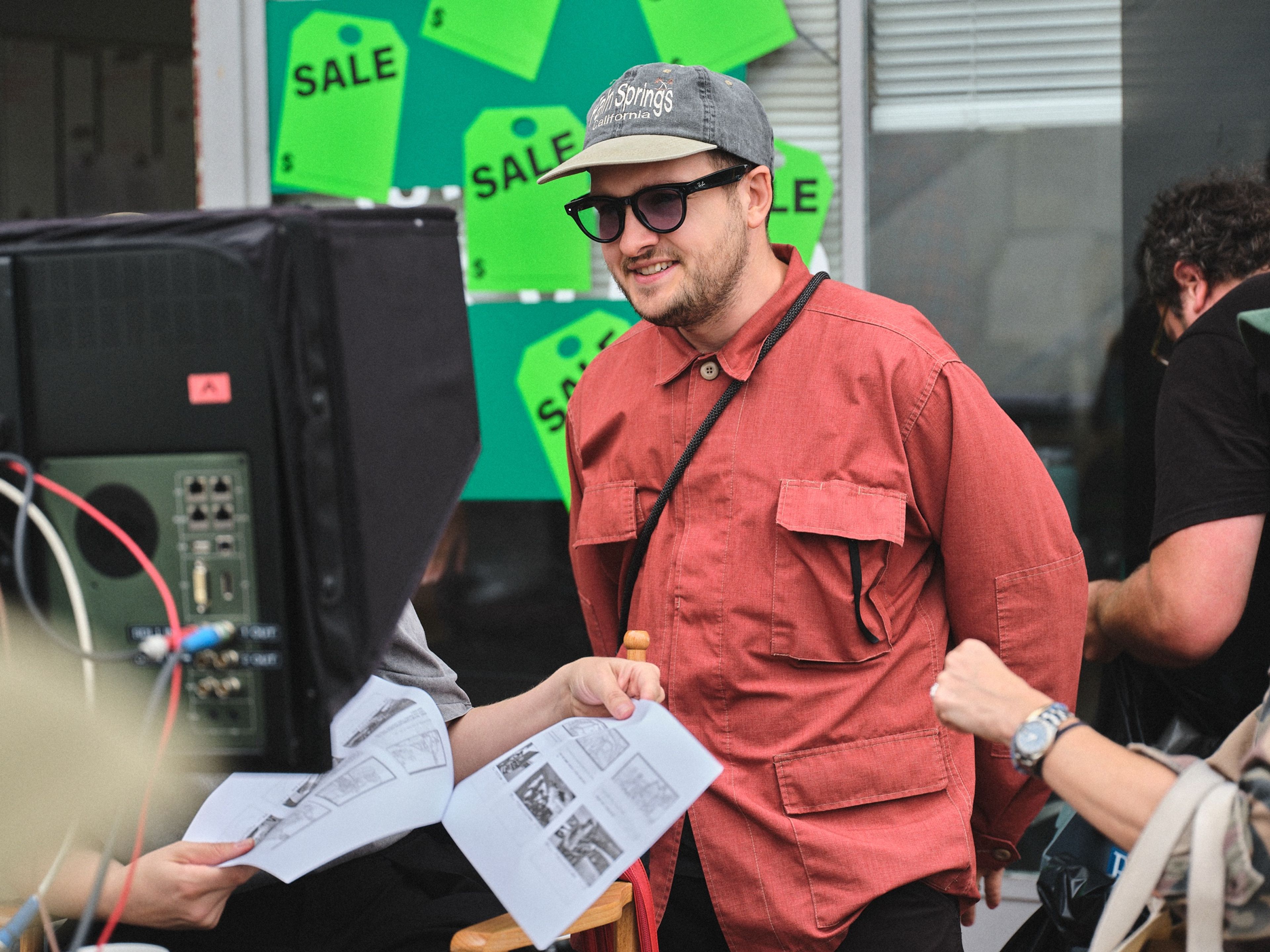 A smiling man in a cap, sunglasses and red jacket stands beside a monitor as someone hands him a page of storyboards.