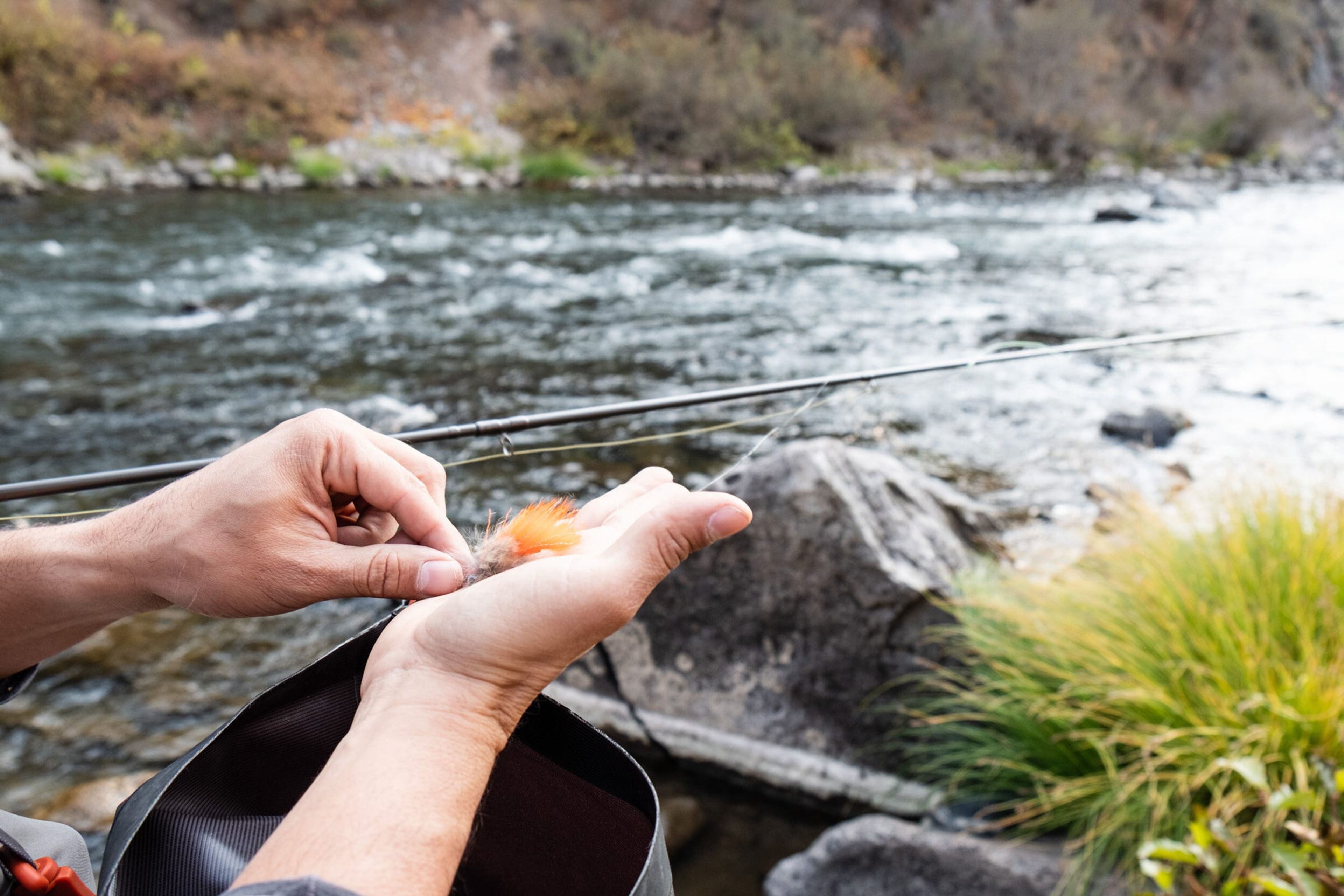 Hands tying an orange fly beside the river
