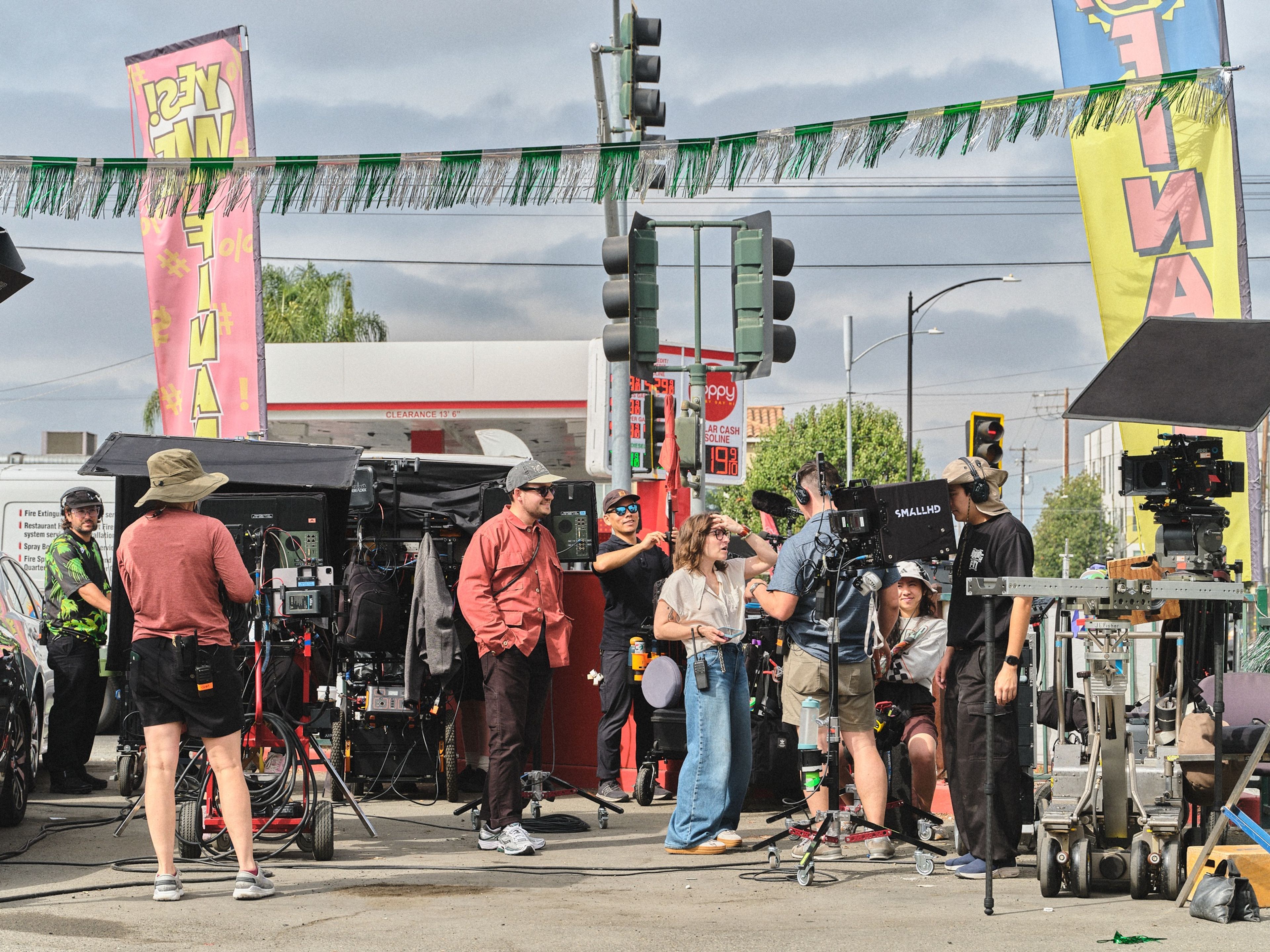 A wide view of the crew gathered by camera carts and monitors at a street corner, under green and silver streamers and tall advertising flags.