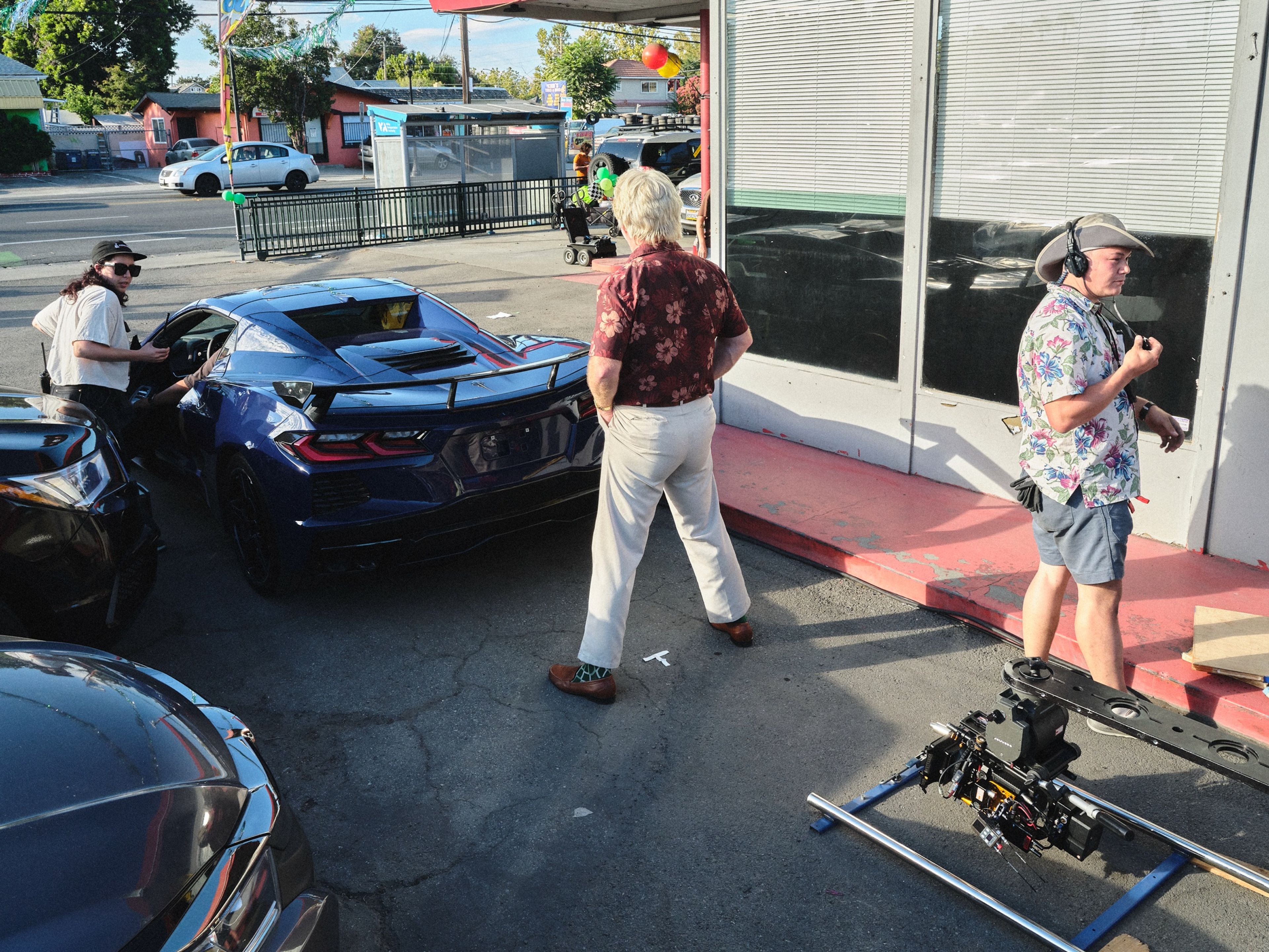 On the car lot, a man with platinum hair in a floral shirt walks toward a blue sports car while a sound mixer in a wide-brimmed hat and headphones stands by a camera slider.