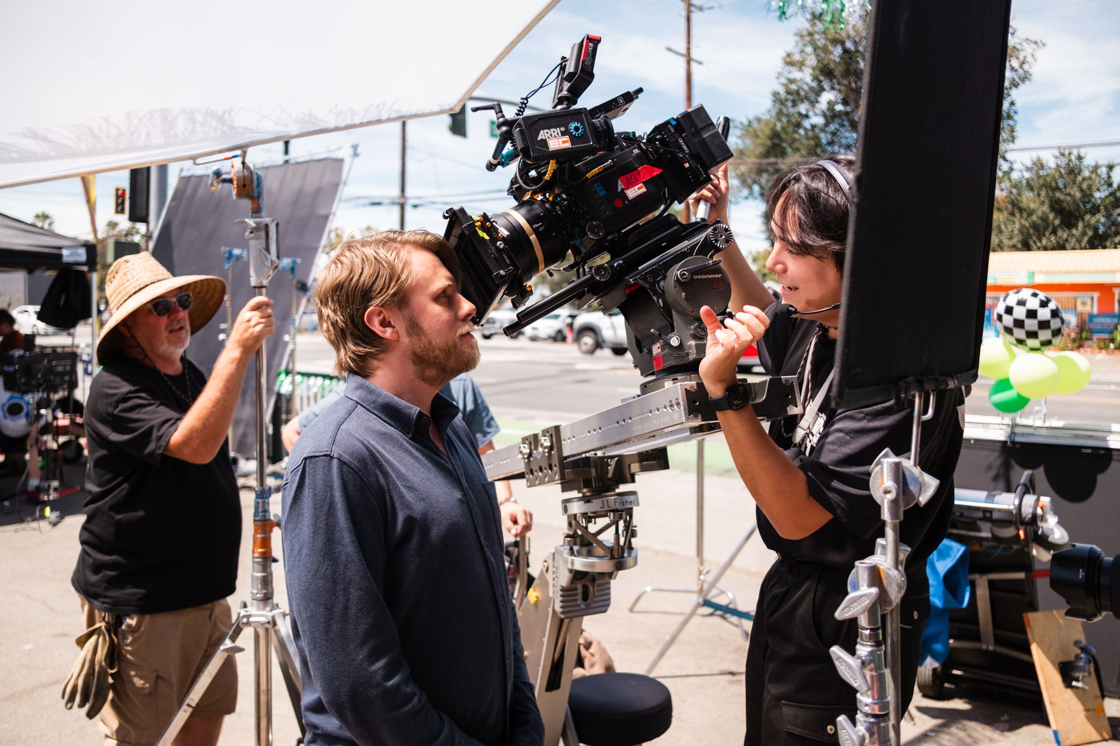 A camera operator adjusts a cinema camera on a slider while a man in a blue shirt looks into the lens and a crew member in a straw hat holds a light stand.