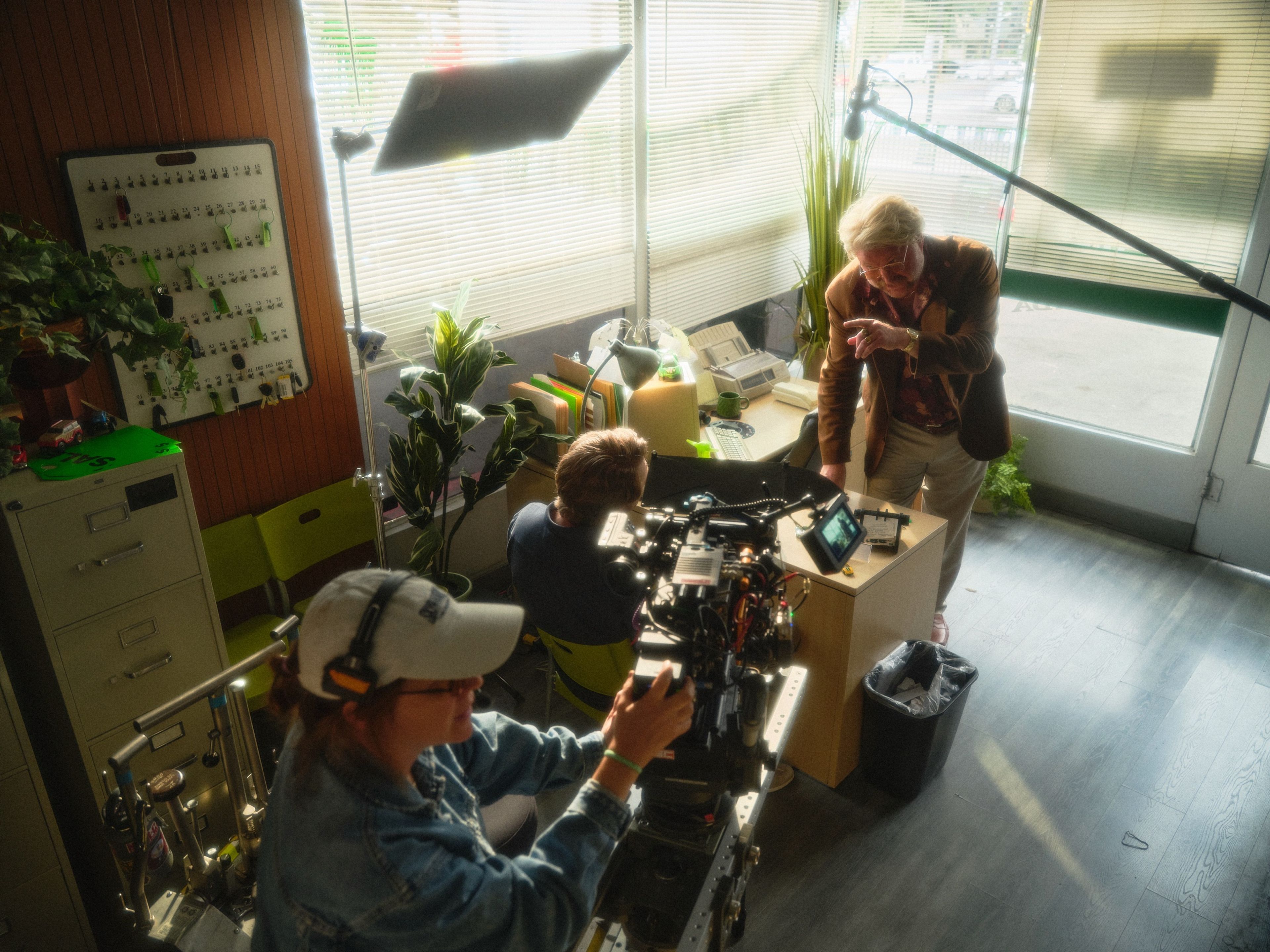 Inside a sales office, a camera operator lines up a shot while an actor in a brown blazer leans over the desk toward a seated actor.