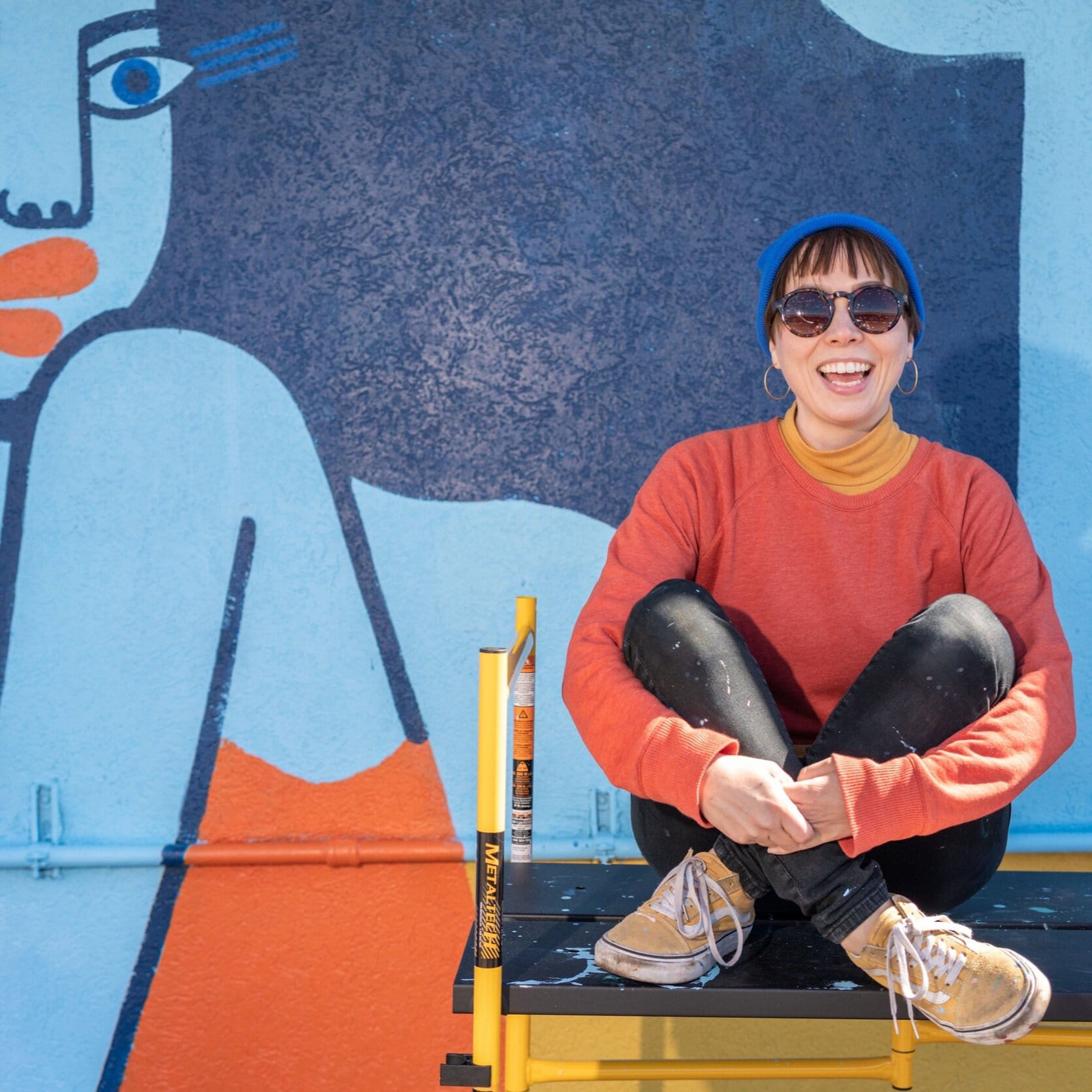 Mural artist in an orange sweater sitting in front of her mural