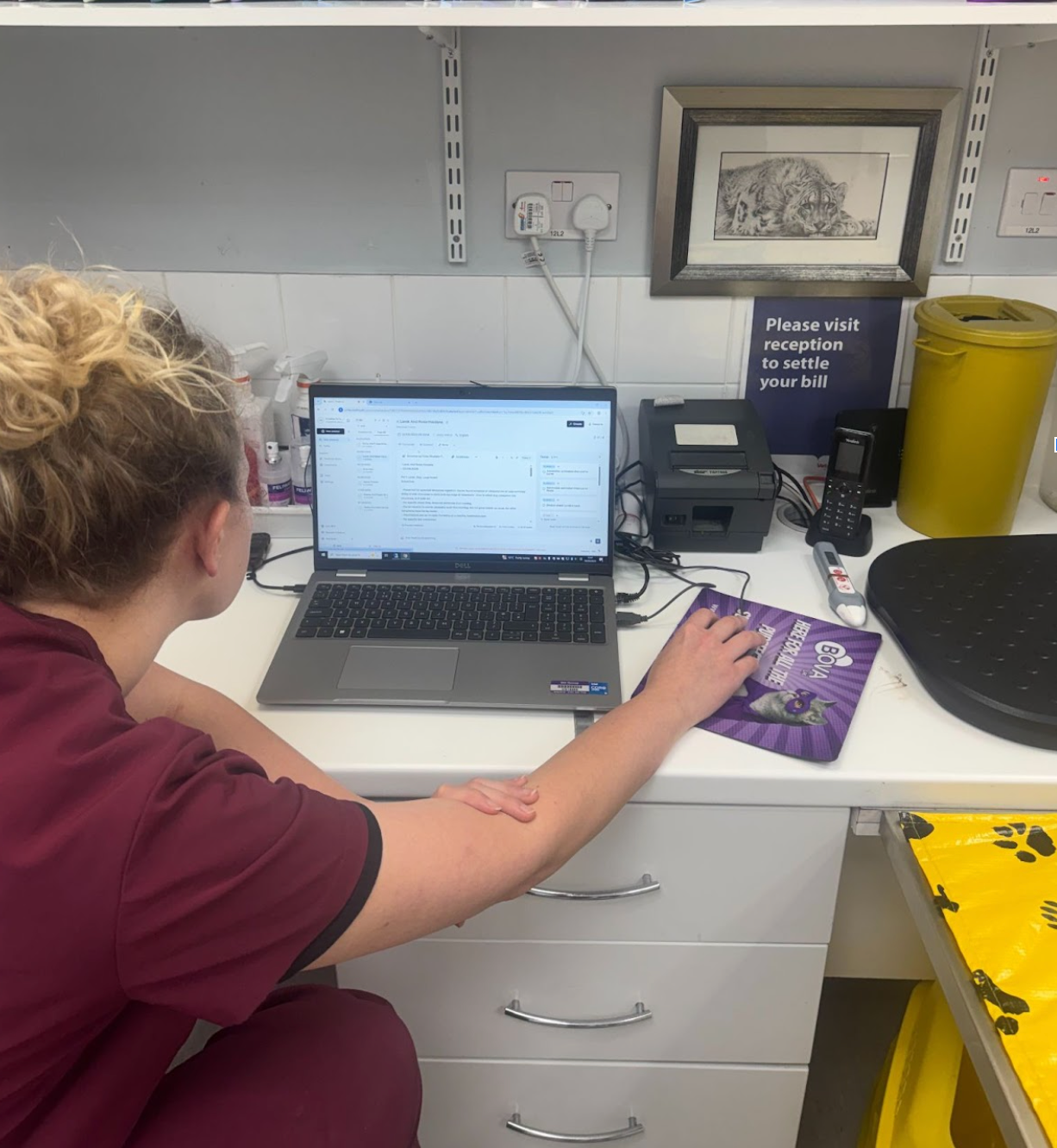 A person in burgundy scrubs uses a laptop and mouse at a desk in a vet clinic reception area.