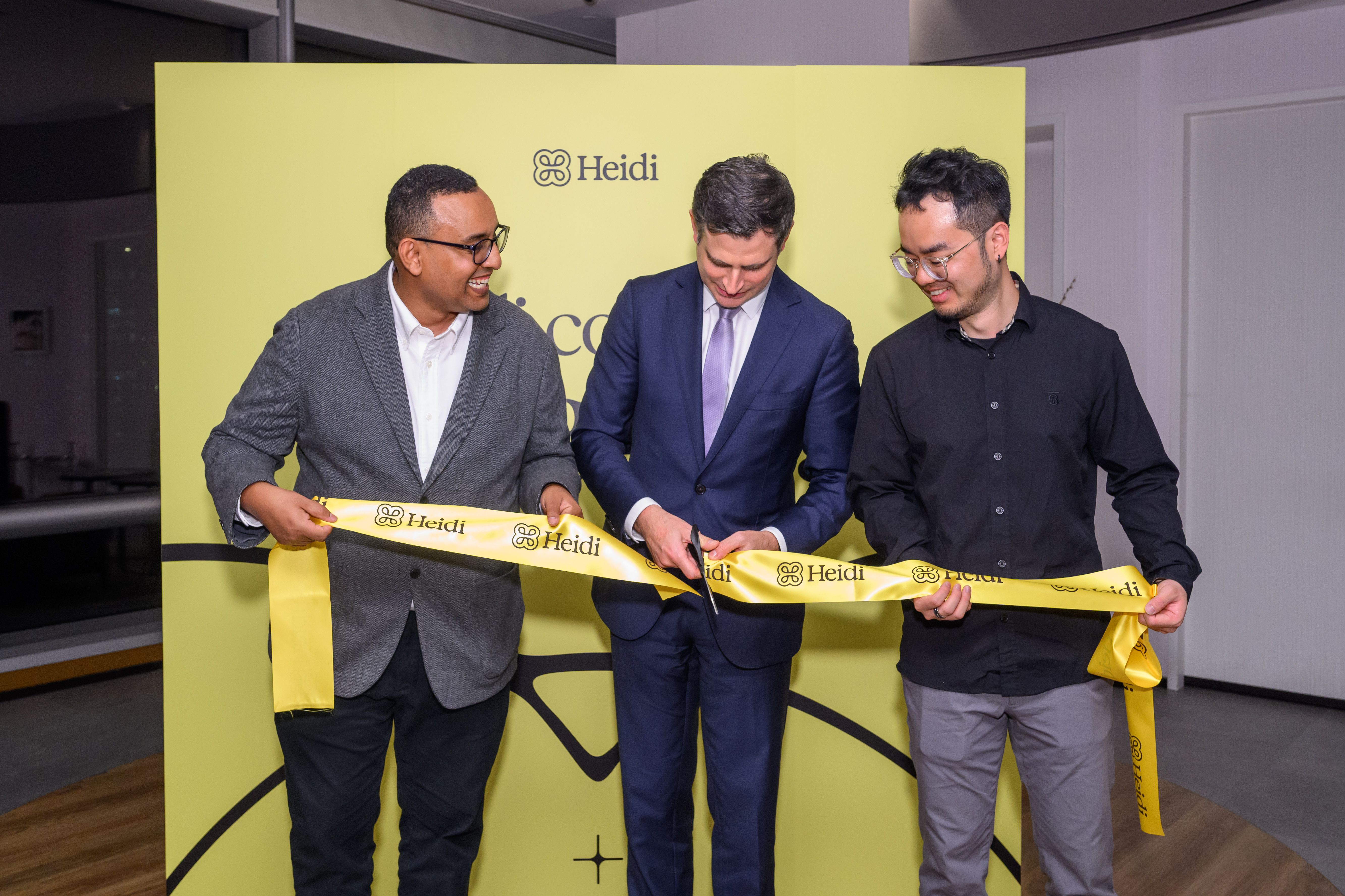 Three men participate in a ribbon-cutting ceremony, with one man in a suit cutting a yellow ribbon branded "Heidi."
