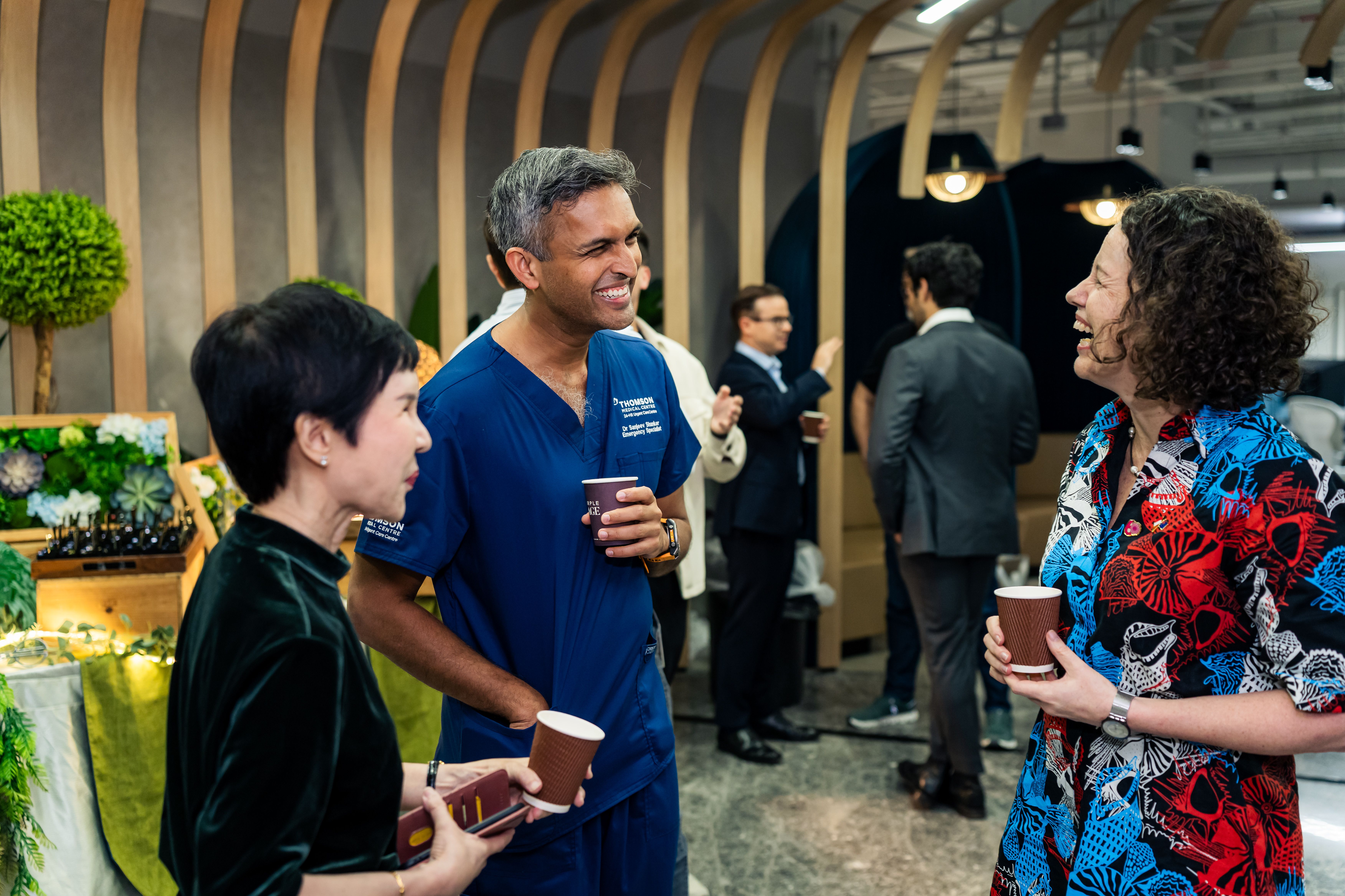 A man in blue scrubs and two women, one in a colorful shirt, smiling and conversing at an event, all holding cups.