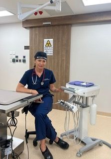A smiling woman in blue scrubs, a headlamp, and a stethoscope sits in a modern medical room with equipment and a radiation warning sign on a door behind her.