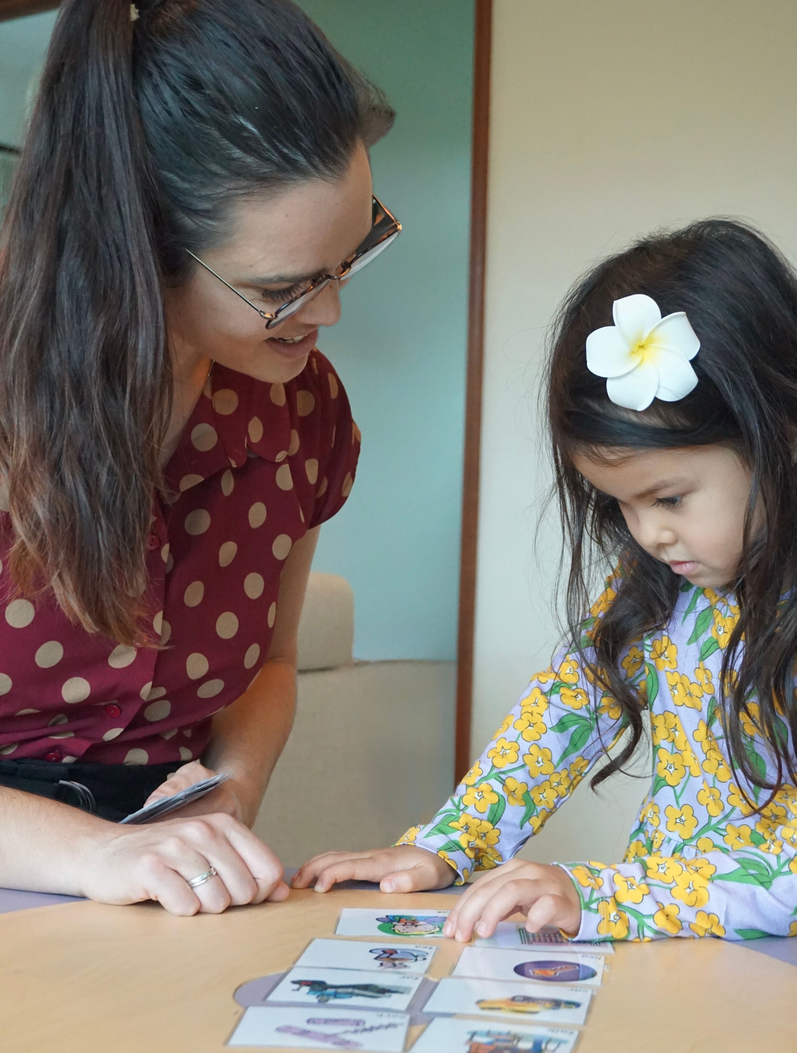 A smiling woman with glasses and a young girl with a flower in her hair look at picture cards on a table.