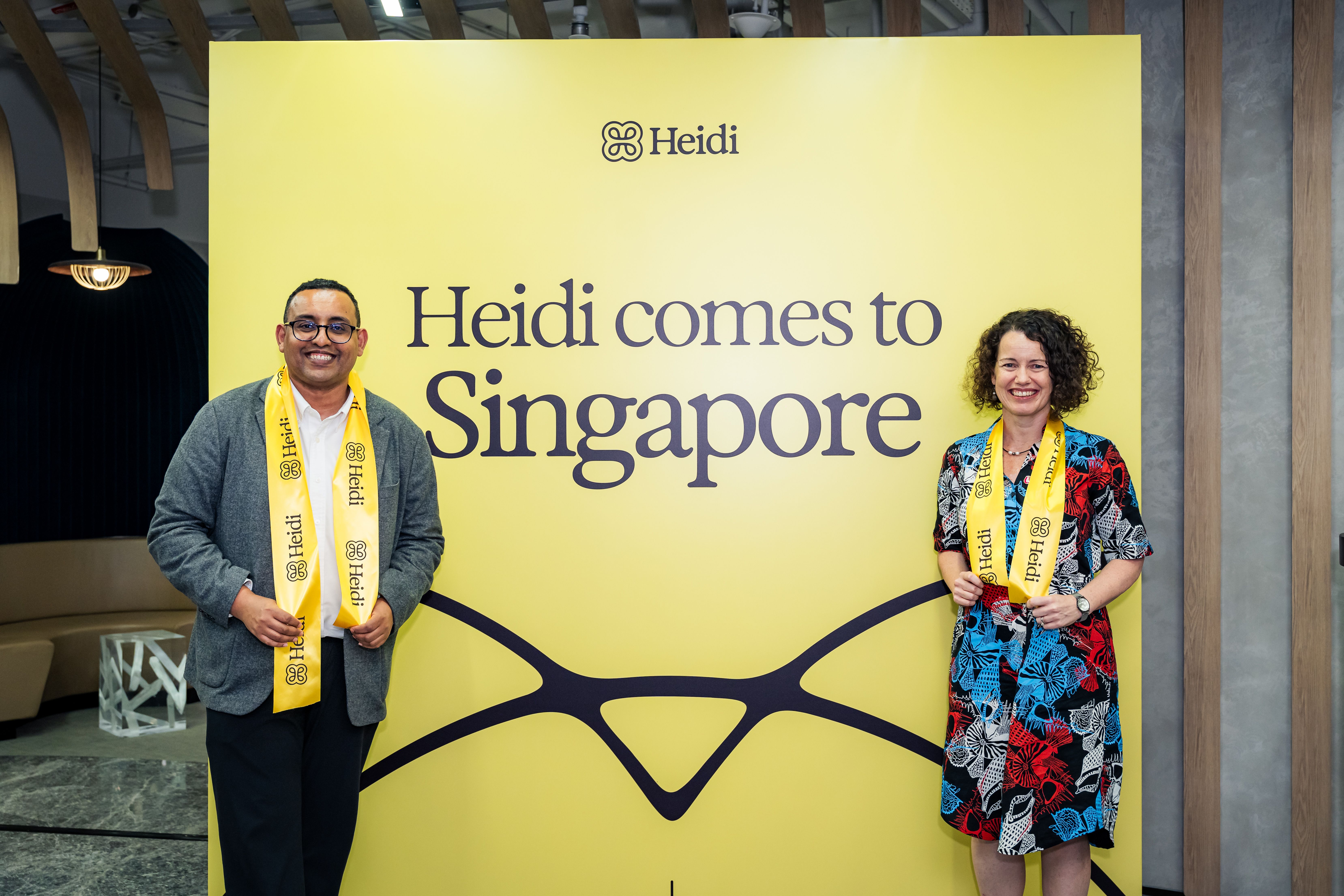 Two people wearing Heidi sashes stand in front of a yellow "Heidi comes to Singapore" banner.