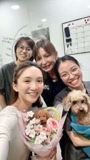 Four smiling women, one holding flowers and a toy dog, another holding a real dog wrapped in a towel.