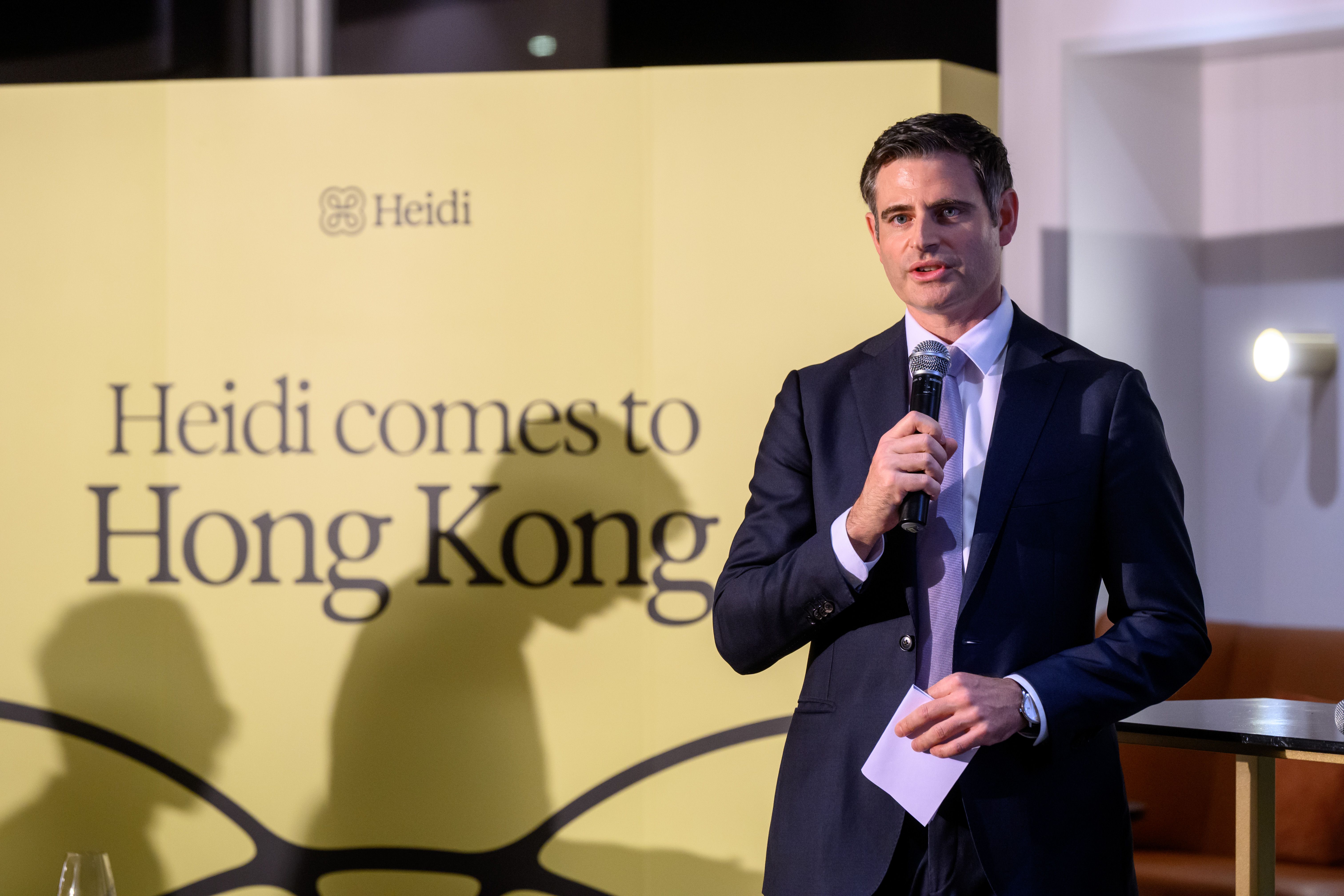 Gareth Williams, Australian Consulate-General to Hong Kong in a suit speaks into a microphone in front of a yellow banner that reads "Heidi comes to Hong Kong."