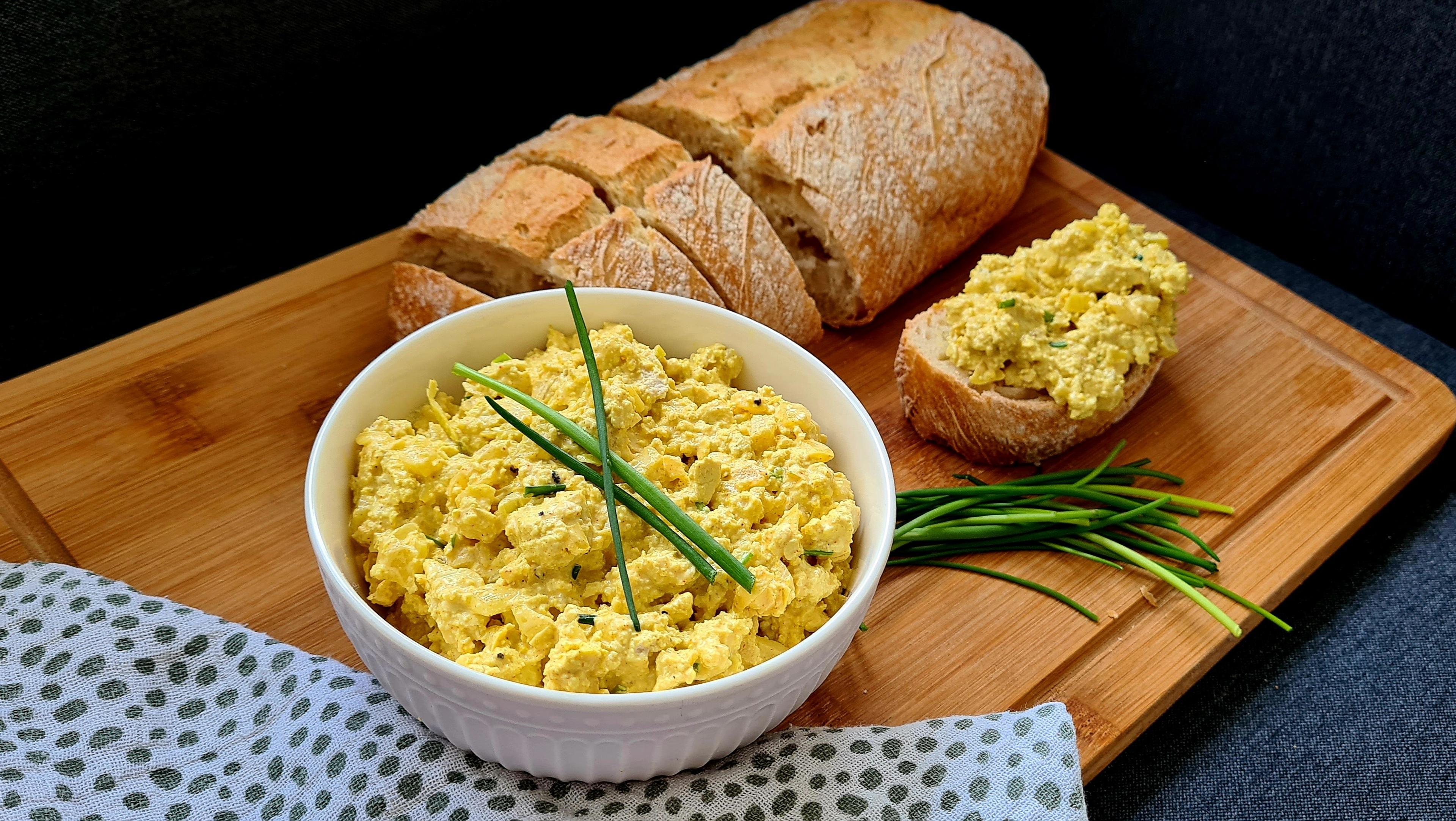 Vegan eiersalade in witte kom naast sneetjes brood op houten serveerplank.
