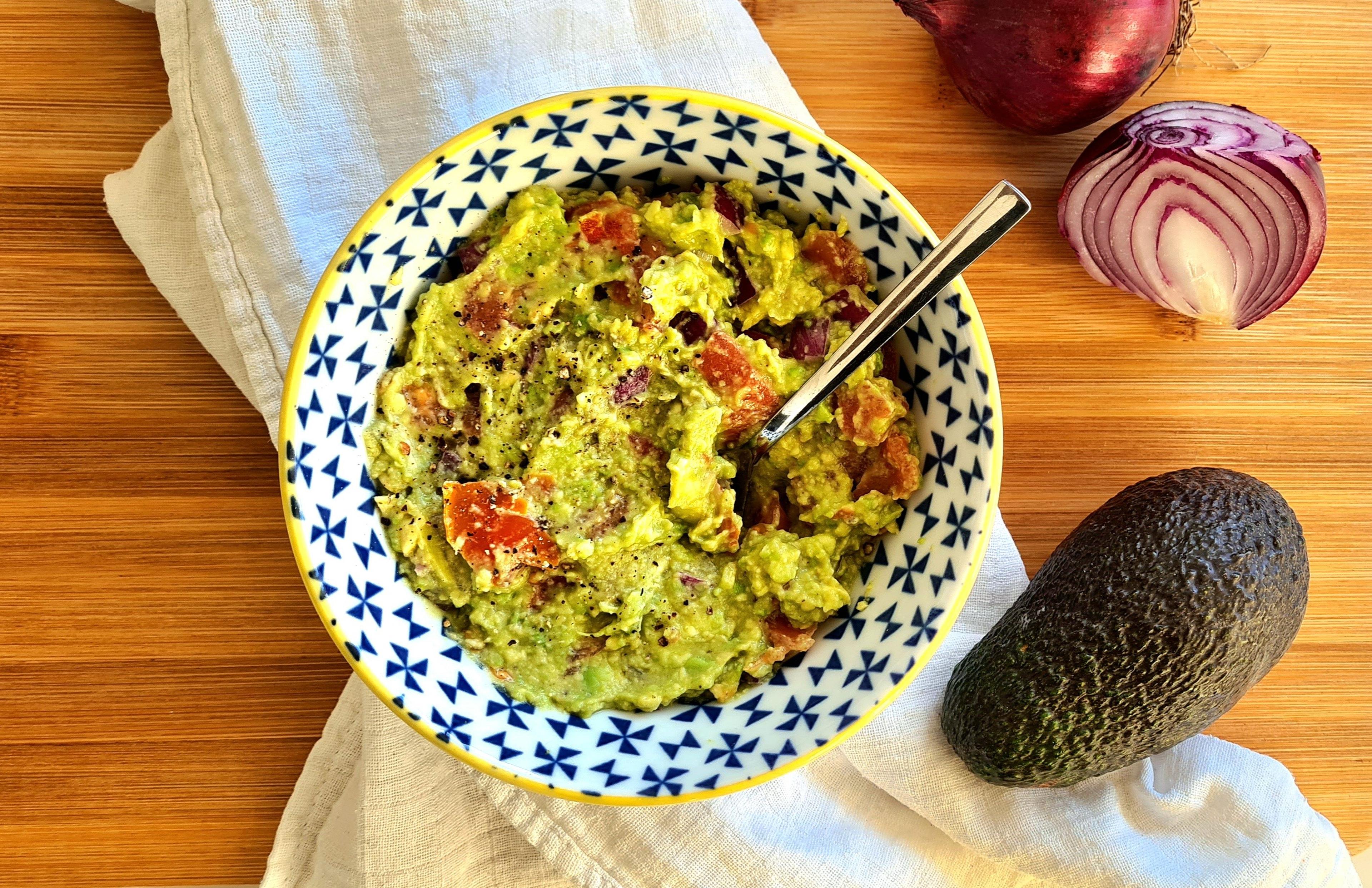 Guacamole in kom naast een avocado en rode ui.