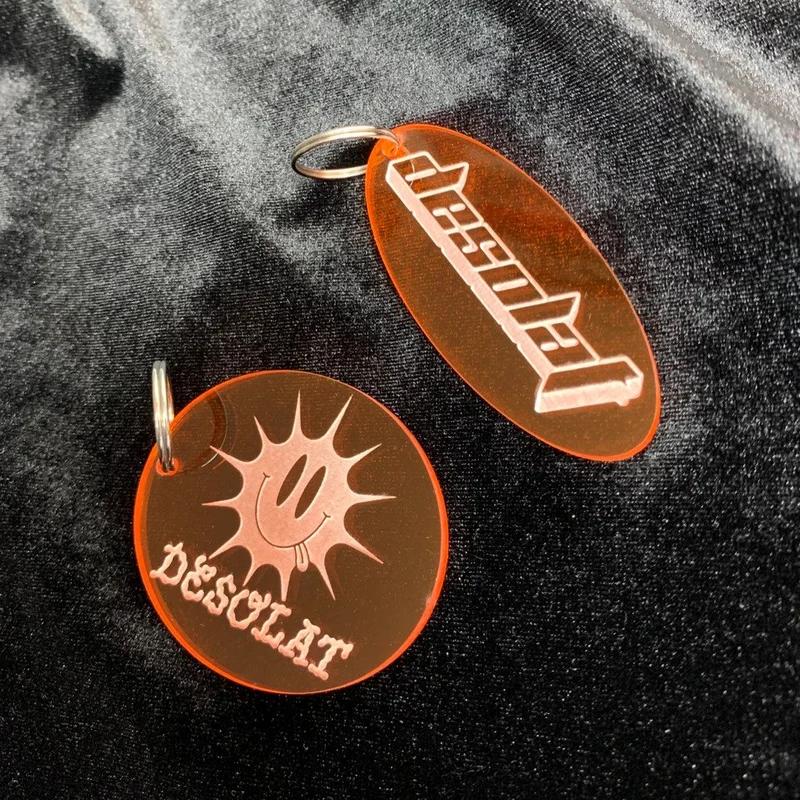 2 Keychain variants in transparent orange, one with a smiling sun logo and the other with main DESOLAT logo.