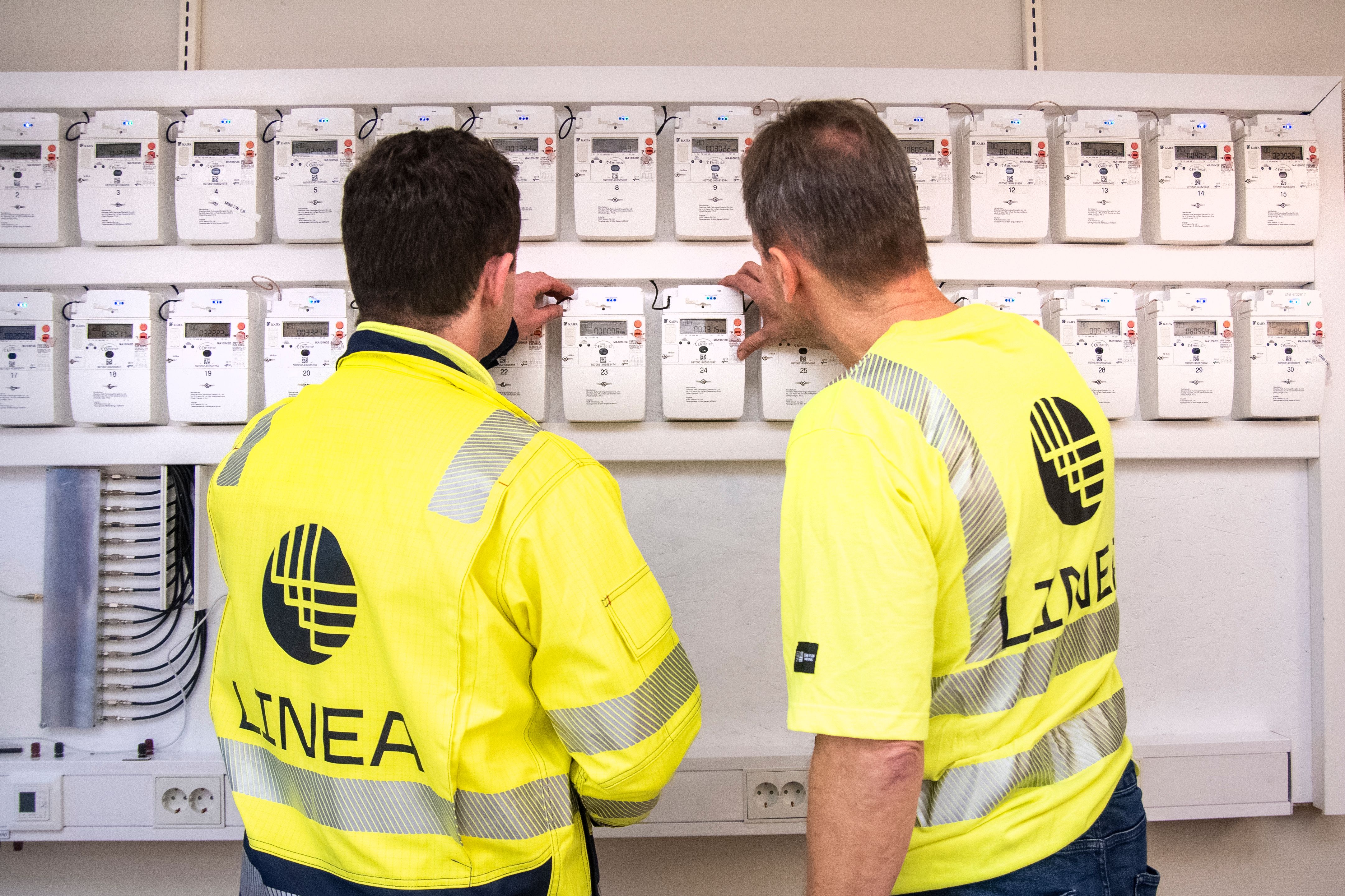 Montører som står å ser på flere strømmålere som henger på vegg.