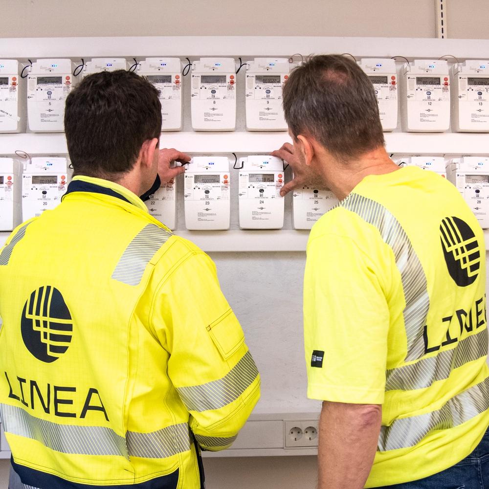 Montører som står å ser på flere strømmålere som henger på vegg.