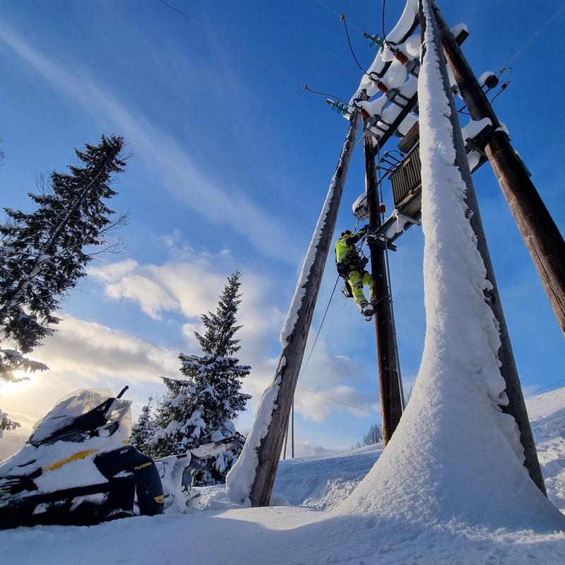 Montør som jobber i stolpe på vinteren, men blå himmel og trær i bakgrunnen.