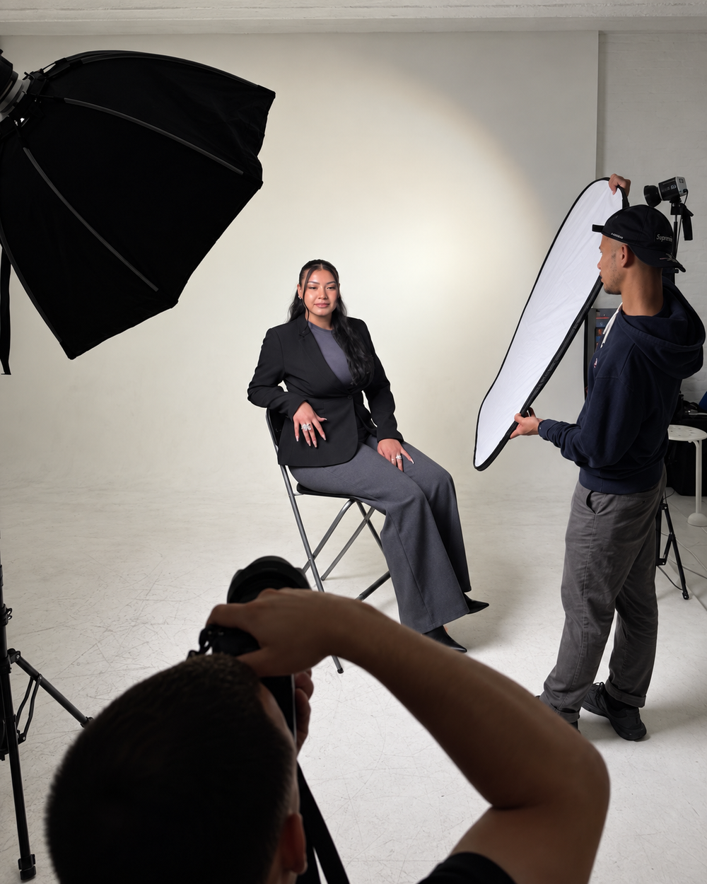 En kvinne sitter foran kamera i et fotostudio mens en assistent holder en reflektor og en fotograf tar bilde.