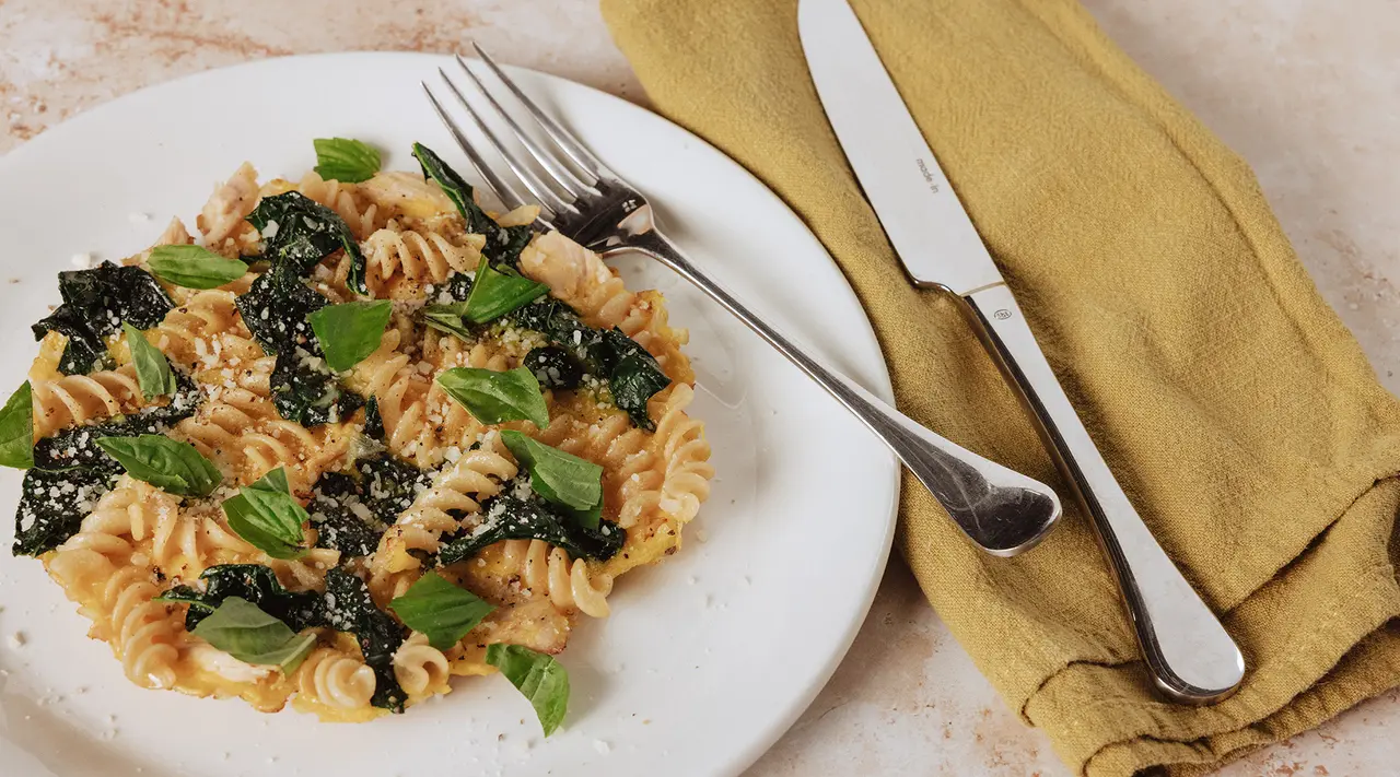 A plate of pasta featuring spiral noodles, topped with greens and garnished with herbs, is accompanied by a fork and knife on a yellow napkin.