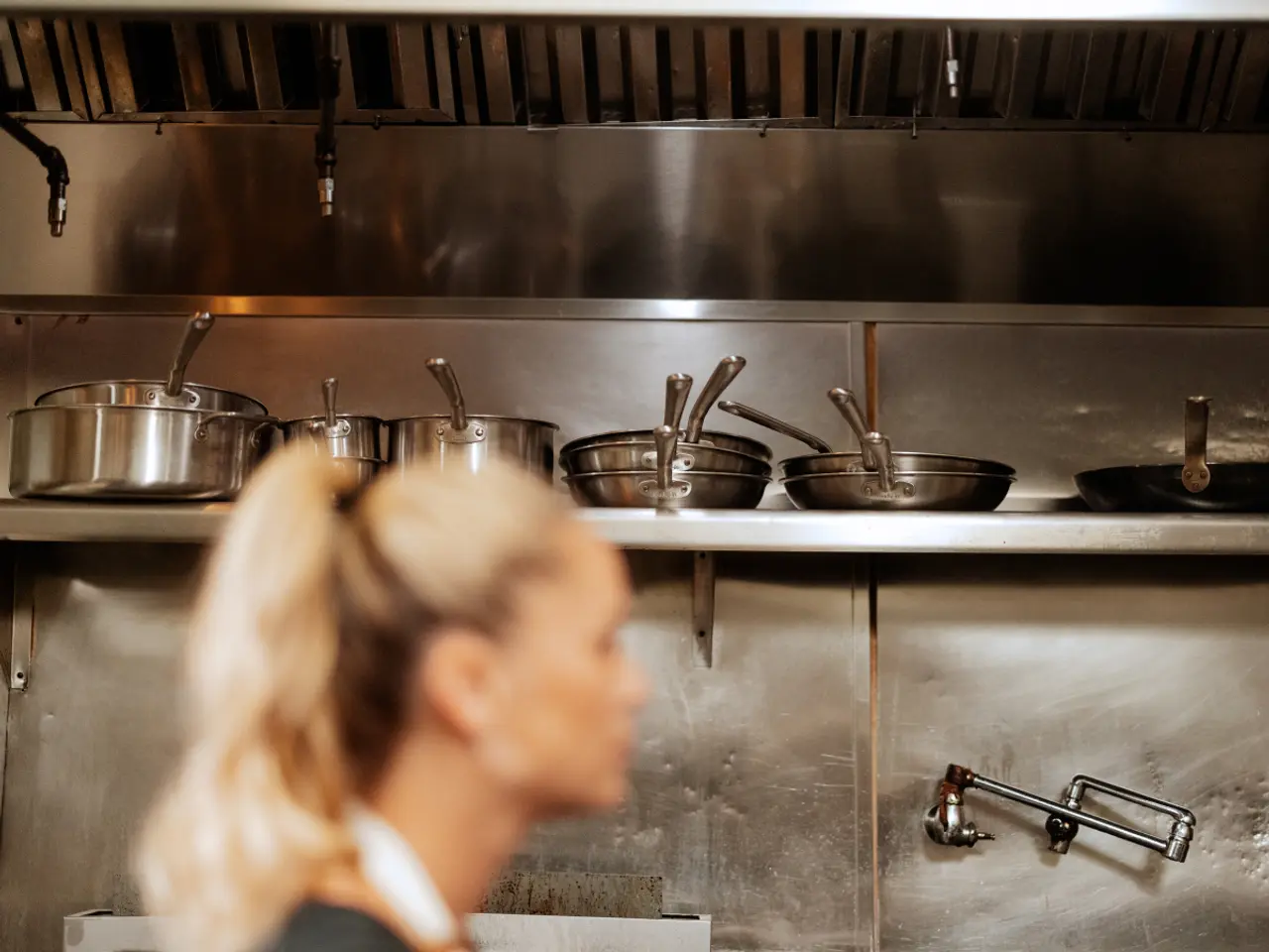 A professional kitchen with stainless steel equipment and cookware is blurred in the background behind a focused woman with her hair tied back.