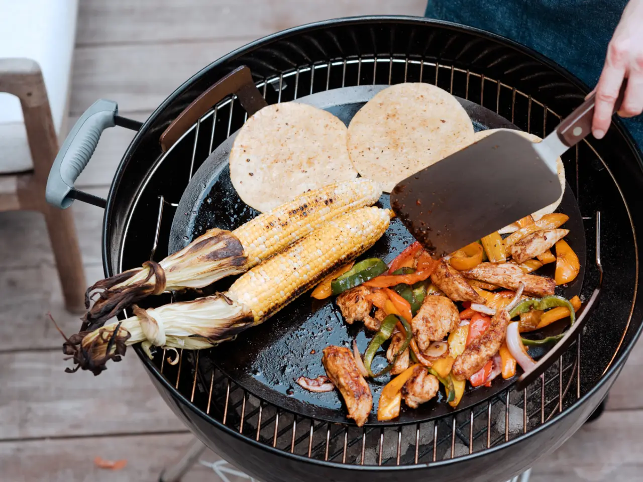 A person grills an assortment of food including corn, tortillas, and mixed vegetables on an outdoor grill.