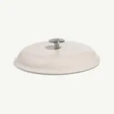 A round, beige cooking lid with a stainless steel handle sits on a light background.