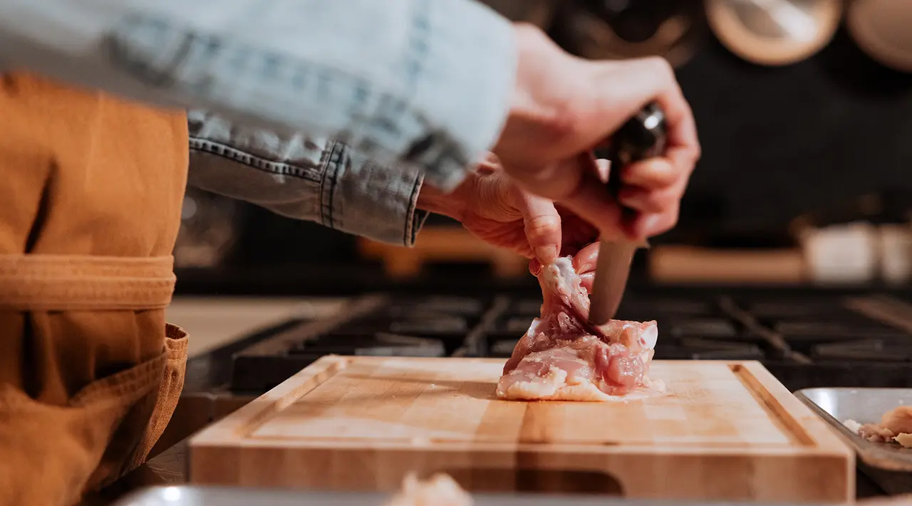 A person is trimming fat from a piece of meat on a wooden cutting board in the kitchen.