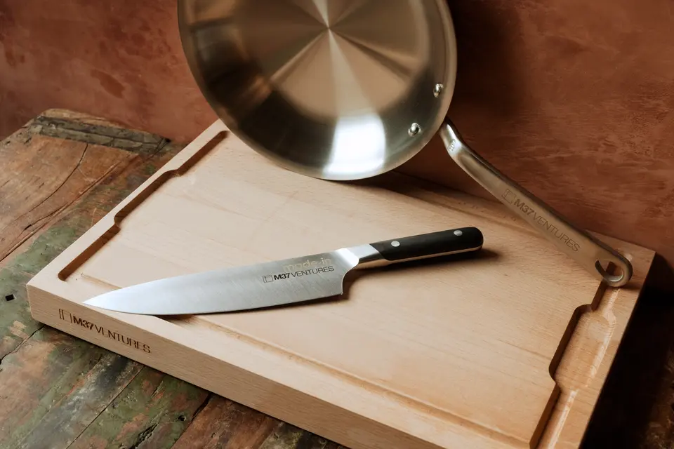 A chef's knife lying on a wooden cutting board next to a stainless steel frying pan on a rustic table.