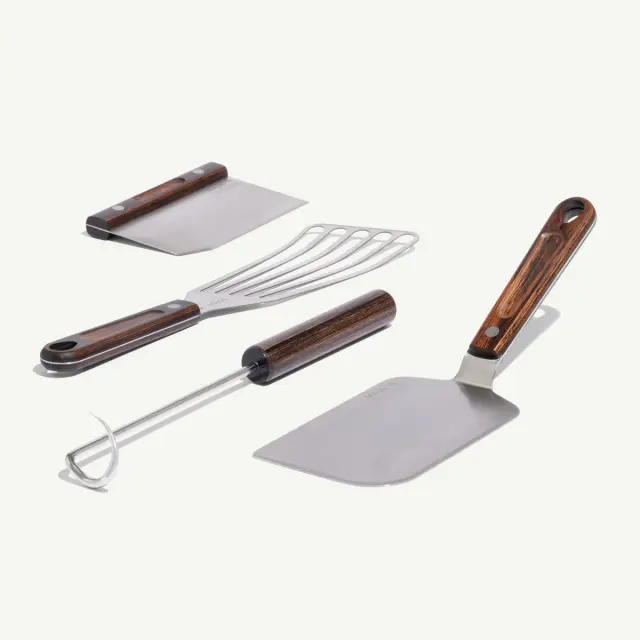 Four stainless steel cooking utensils with wooden handles, including a spatula, grill fork, dough cutter, and a slotted turner, arranged neatly.