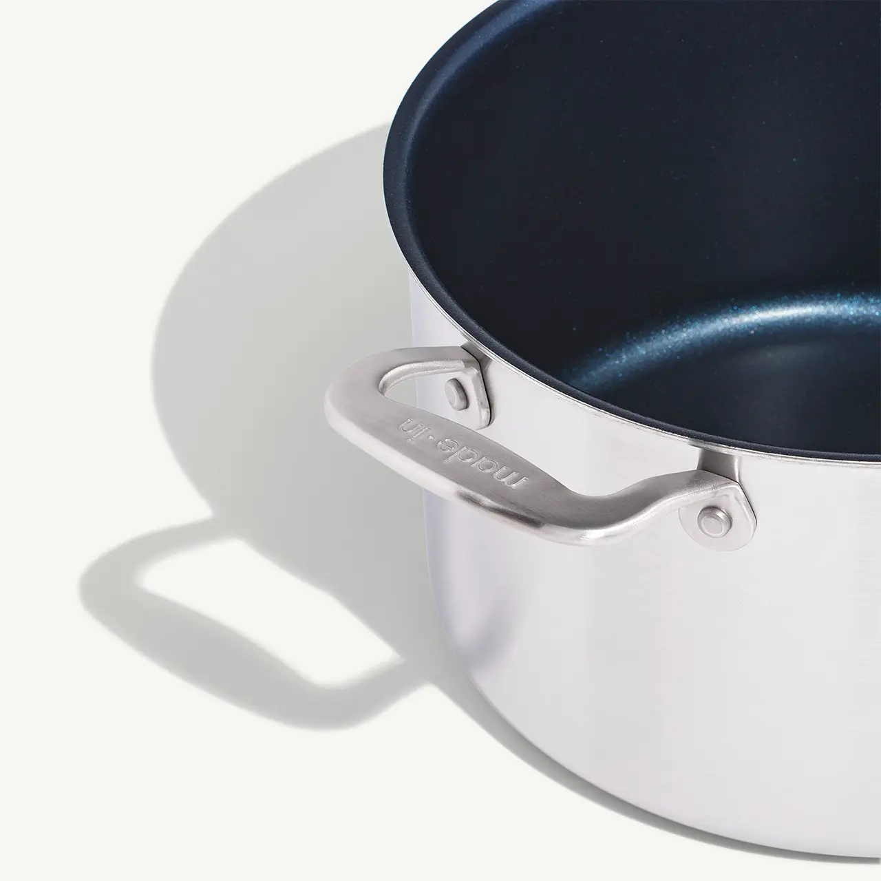 A stainless steel pot with a blue interior casts a shadow on a white surface, highlighting its shiny handle with the word "Induction" engraved on it.