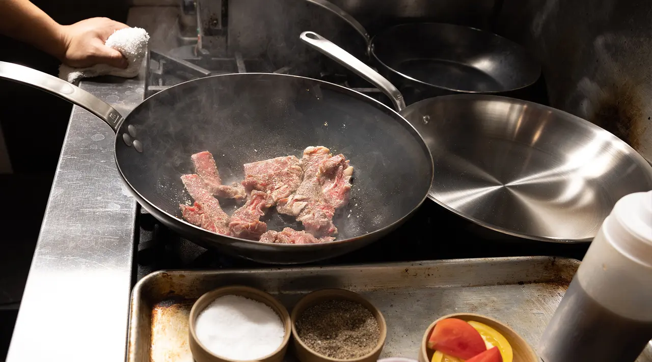 A chef's hand is visible tossing slices of meat in a wok on a stove, with spices and ingredients nearby, indicating a meal being prepared.