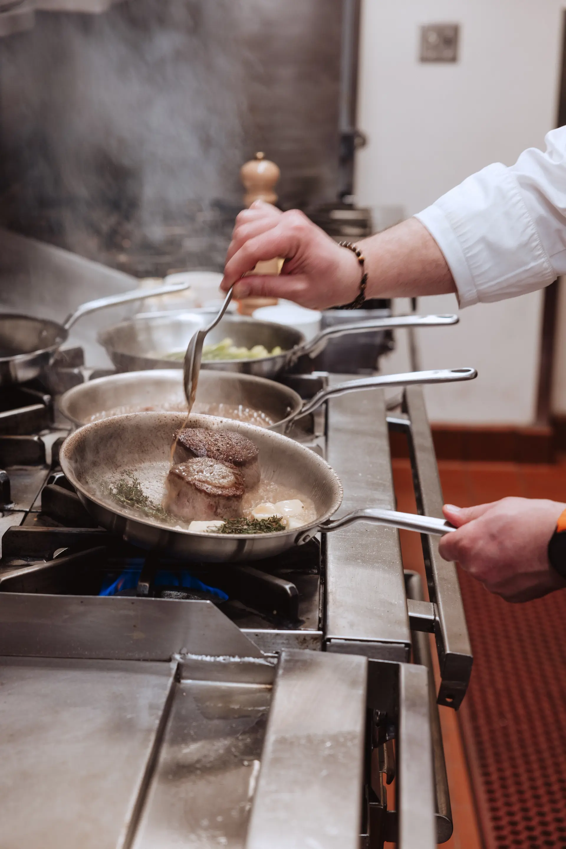 A chef is sautéing meat in a pan over a gas stove in a busy kitchen environment.