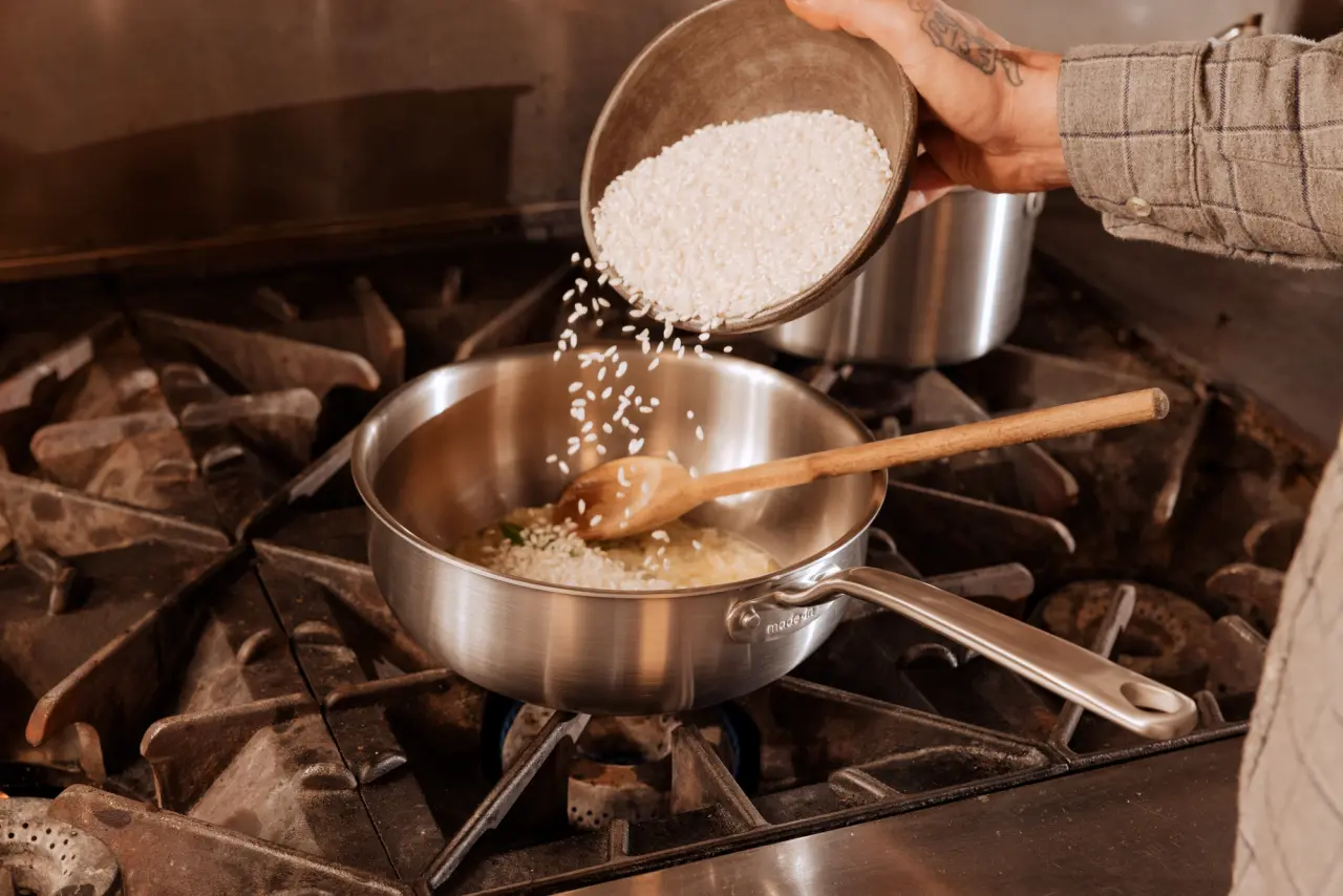 A person is pouring rice into a stainless steel saucepan on a gas stove while stirring with a wooden spoon.