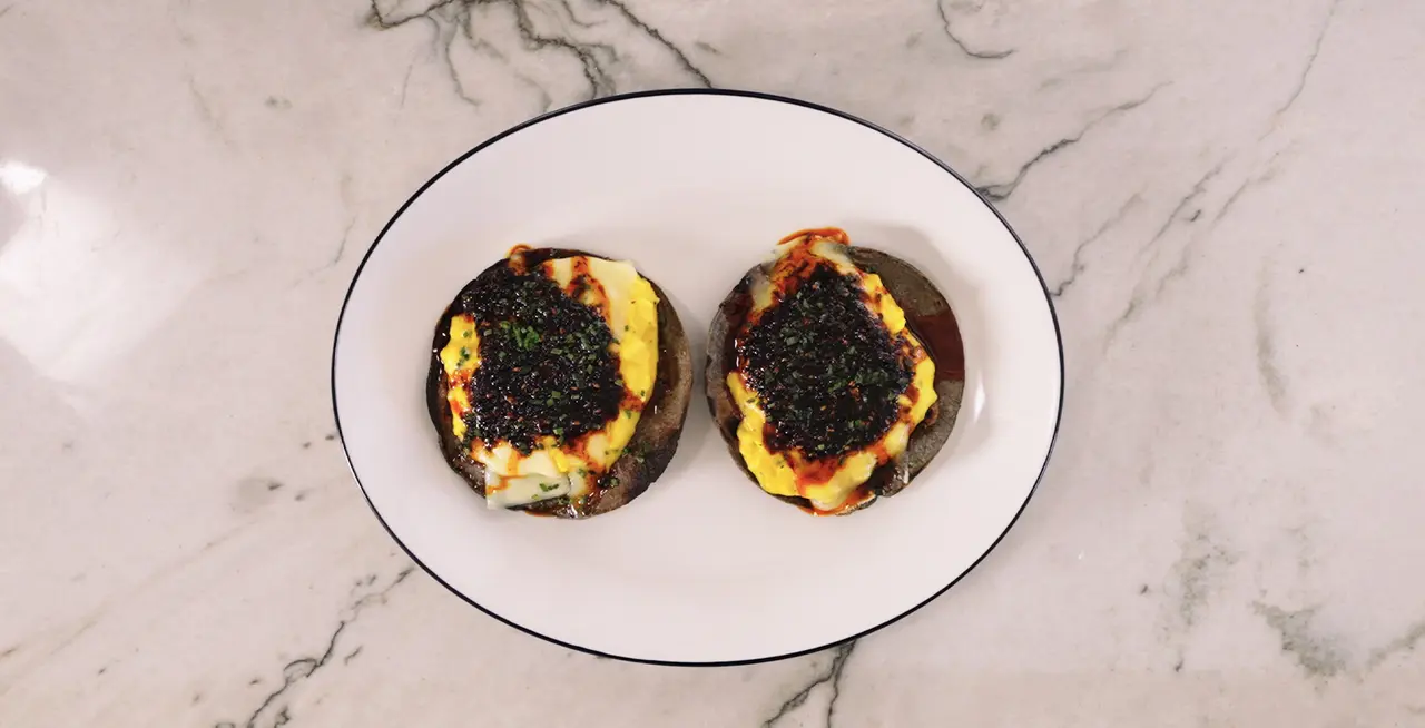 Two English muffin halves topped with cheese, sauce, and black caviar are presented on a white plate against a marble countertop.