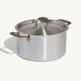 A stainless steel cooking pot with a lid and wooden handles sits on a light background.