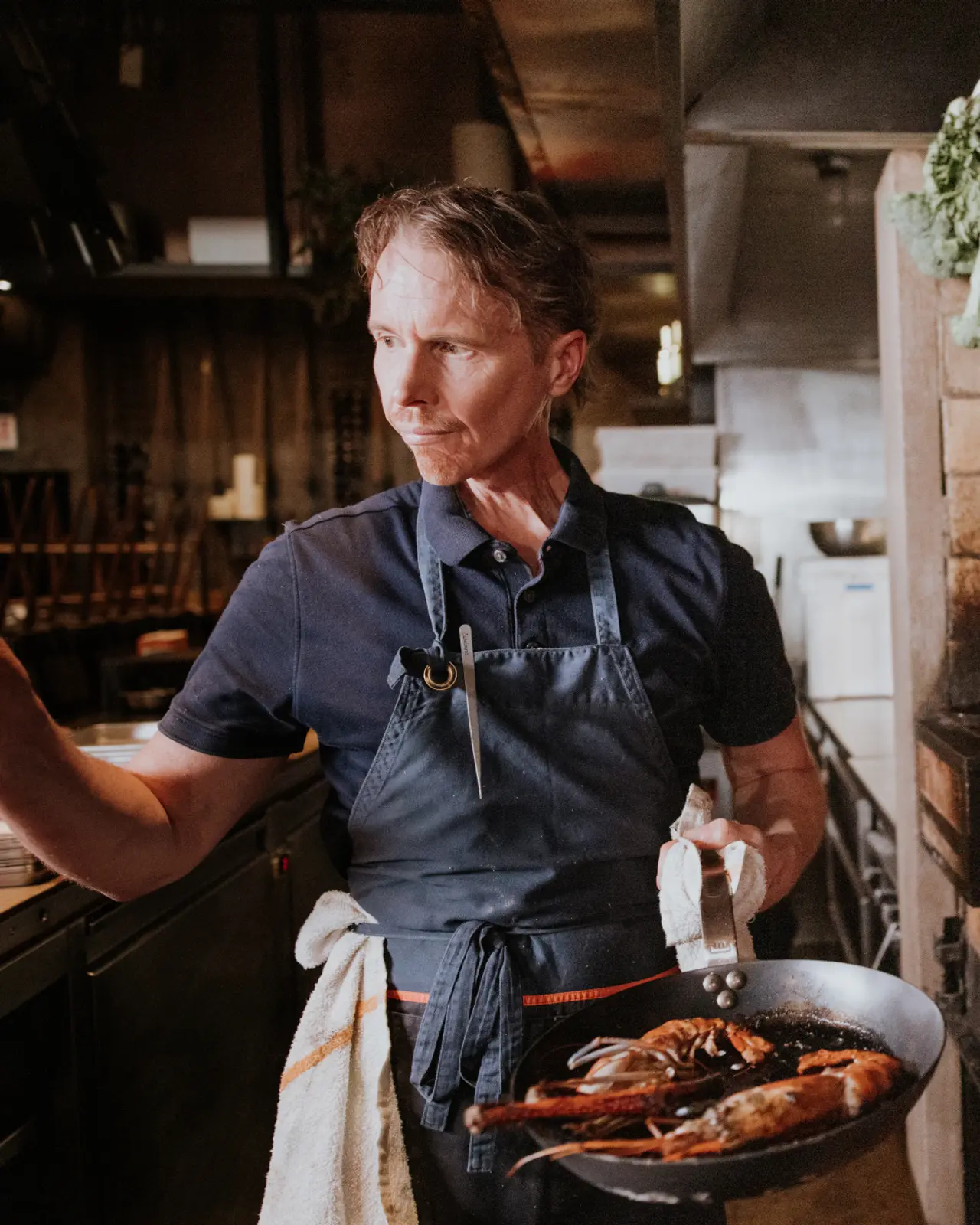 A focused chef holds a pan with lobsters inside a restaurant kitchen.