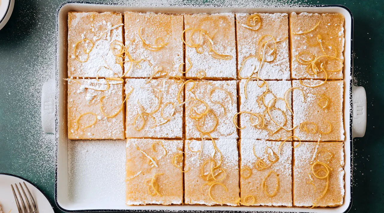 A tray of freshly baked lemon bars is dusted with powdered sugar and garnished with lemon zest.