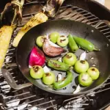 Grilled vegetables, including tomatillos, jalapeños, and red onions, are arranged in a black skillet on a barbecue grill alongside corn on the cob.