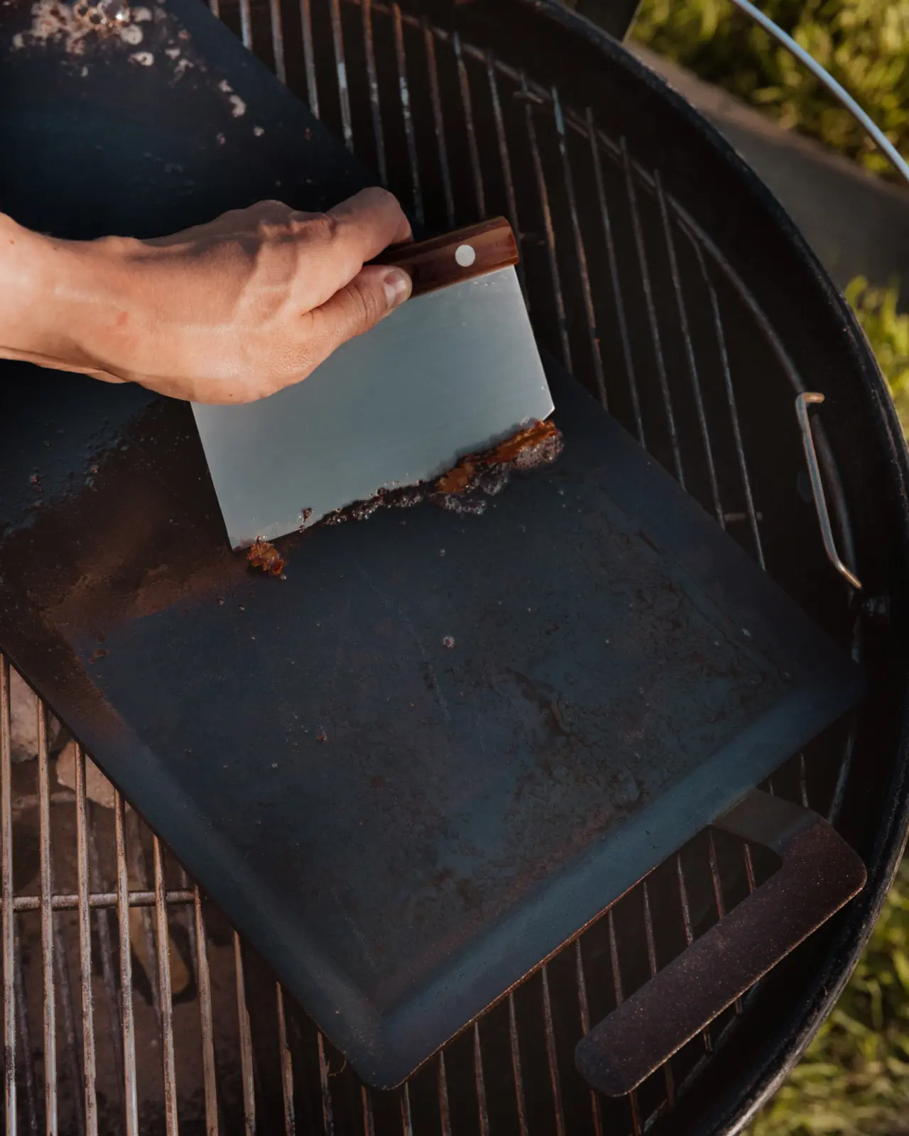 A person's hands are using a scraper to clean a grill, removing charred residue from its surface.