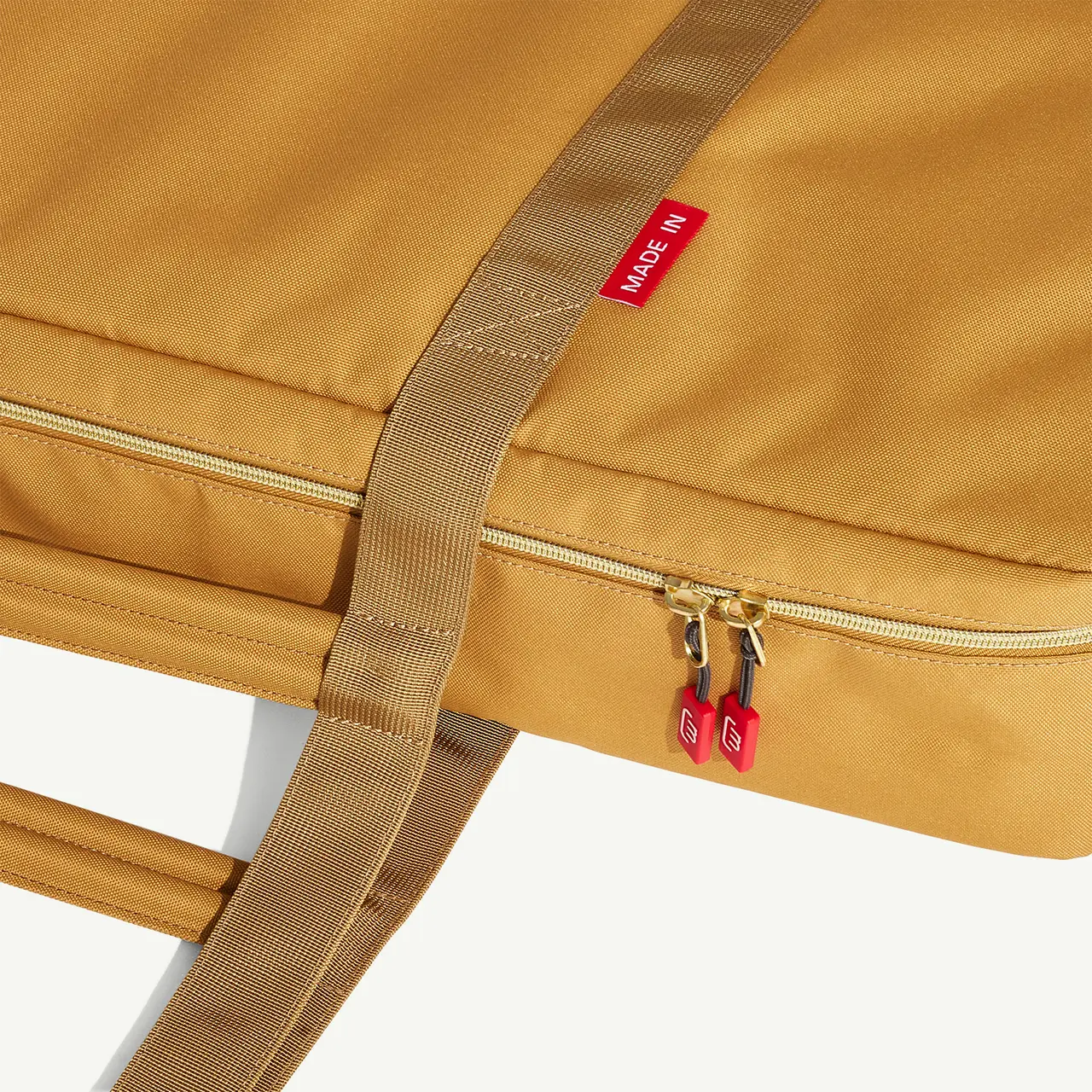 A close-up view of a mustard-yellow backpack featuring a front zip pocket with red zipper pulls and a red brand tag on the side.