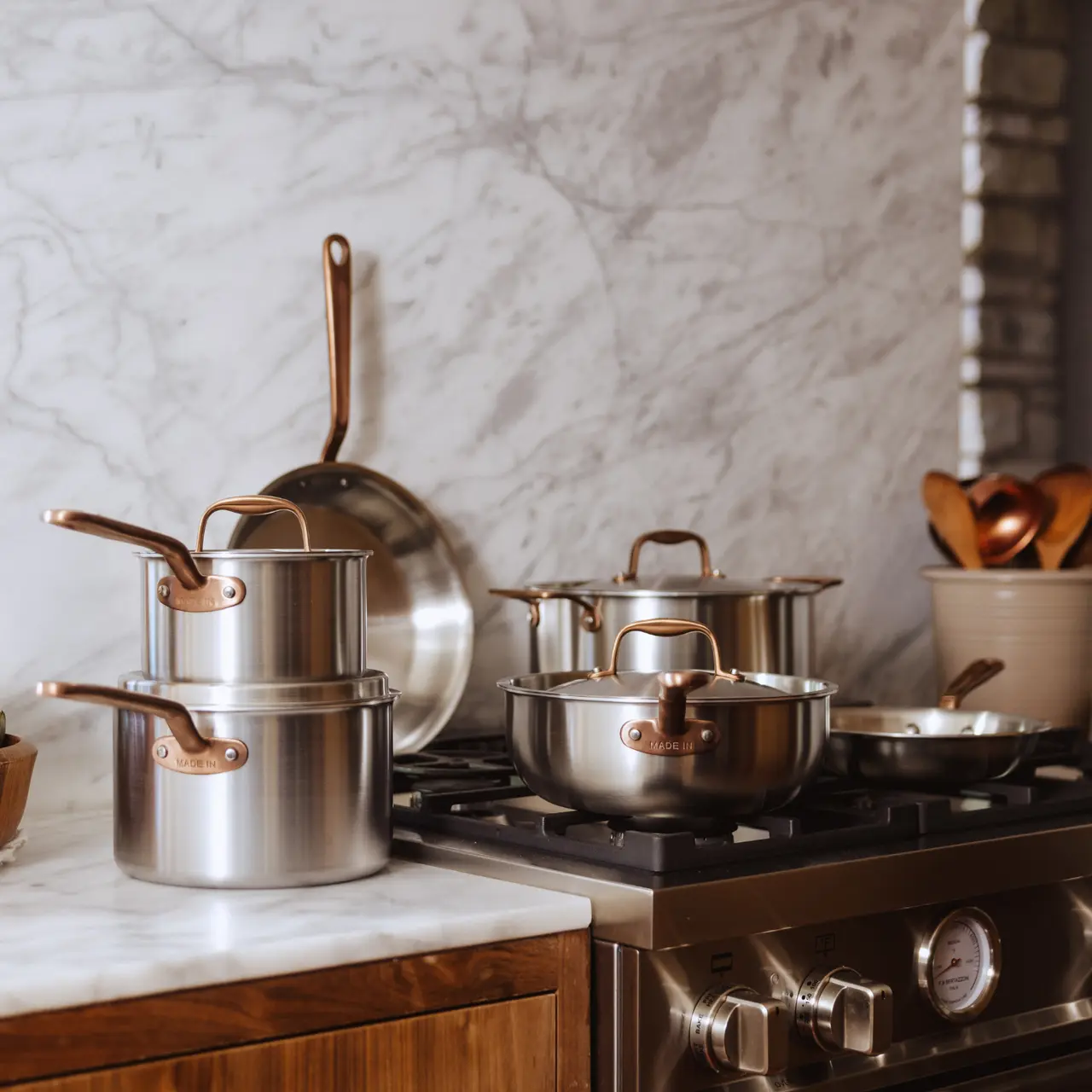 A modern kitchen countertop features an assortment of stainless steel pots and pans with copper handles, alongside wooden utensils and a marble backdrop.