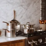 A modern kitchen countertop features an assortment of stainless steel pots and pans with copper handles, alongside wooden utensils and a marble backdrop.