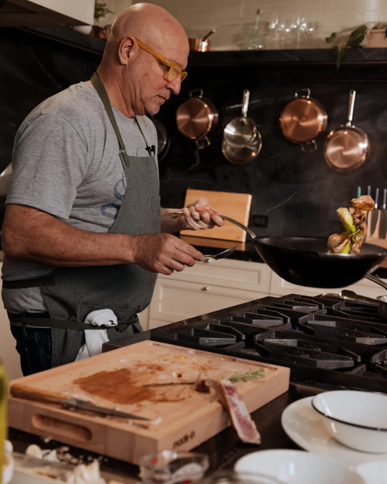 A focused chef wearing glasses and an apron sautés vegetables in a pan in a kitchen with hanging copper pots and a chopping board in the foreground.