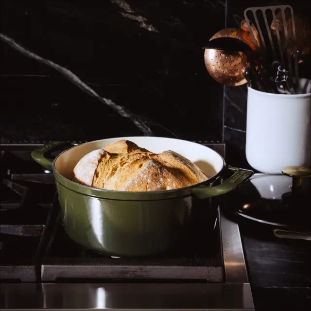 A round loaf of freshly baked bread sits in a green Dutch oven on a black stovetop, with kitchen utensils nearby.